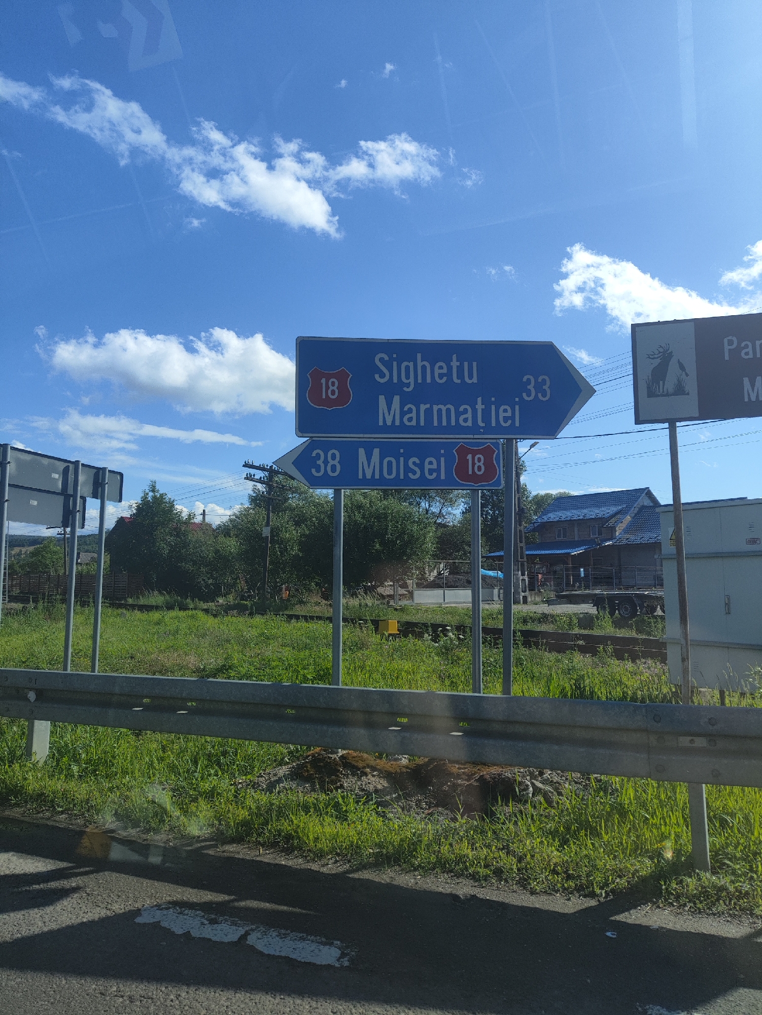 The image captures a road scene under a bright blue sky with scattered white clouds, likely taken from a moving vehicle in Petrova, Romania. Two blue directional signs are prominent. The upper sign, an arrow pointing right, reads "Sighetu Marmatiei" with the number "33". Below this, another arrow pointing right displays "38 Moisei" with the number "18" in a red shield. To the right of the signs, a separate sign features a silhouette of a deer and the partial word "Par". The foreground is dominated by a metal guardrail and a grassy embankment. Beyond the signs, lush green vegetation and trees are visible, with a glimpse of a building with a blue roof and a trailer behind it. Power lines stretch across the scene, supported by wooden poles. The road surface in the very bottom of the image shows some white markings.