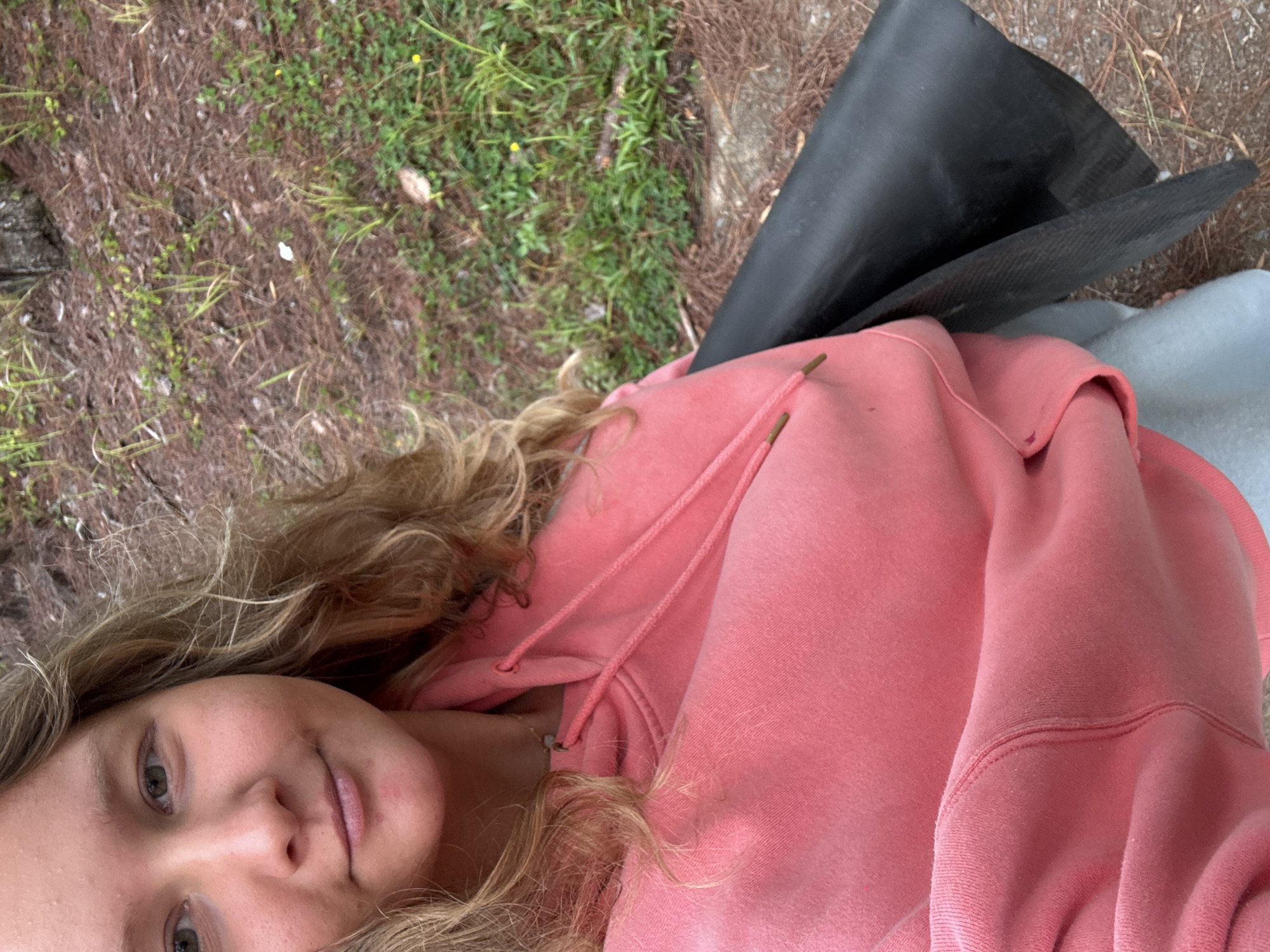 The image shows a close-up of a young woman wearing a pink hoodie. She has blonde, wavy hair and fair skin. Her left eye is visible, along with her nose and mouth. A small, light-colored pendant is visible on a necklace around her neck. Her hoodie has drawstrings with wooden tips. To her right, a black object is partially visible, which appears to be a roll of material, possibly fabric or plastic sheeting. The background is out of focus, but it appears to be an outdoor environment with dry grass, pine needles, and dirt. There are some small green plants and yellow wildflowers visible. The lighting suggests it is daytime. There is no specific information that can place this image in Lac Duong, Vietnam.