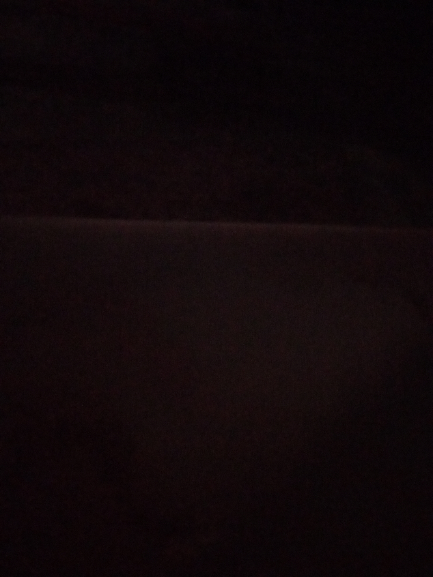 The image is a dark, blurry photograph with a horizontal line dividing the frame approximately in the middle. The upper portion of the image is uniformly dark, while the lower portion is slightly lighter but still predominantly dark. There are no discernible subjects, objects, people, or actions visible in the image. The scene appears to be an interior or a poorly lit outdoor environment with a focus on a textural surface. The location in Lhokseumawe, Indonesia cannot be determined from the visual information provided.