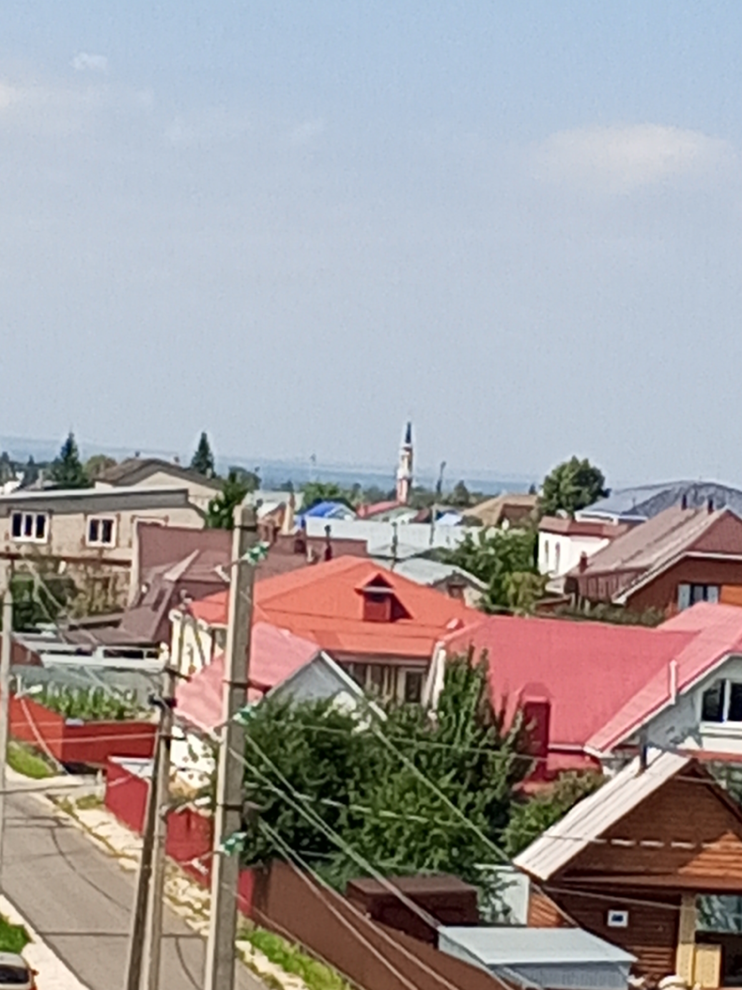 An elevated, clear, sunny, daytime, medium-shot captures a residential area in Chistopol, Russia. The sky is mostly clear with a few wispy clouds. Several houses with red and brown roofs are visible, interspersed with green trees and a street. A tall, slender white tower with a dark pointed top, possibly a church steeple, stands in the distance, to the left of center. Power lines and poles are prominent in the foreground and midground. The overall impression is of a small town or village setting.