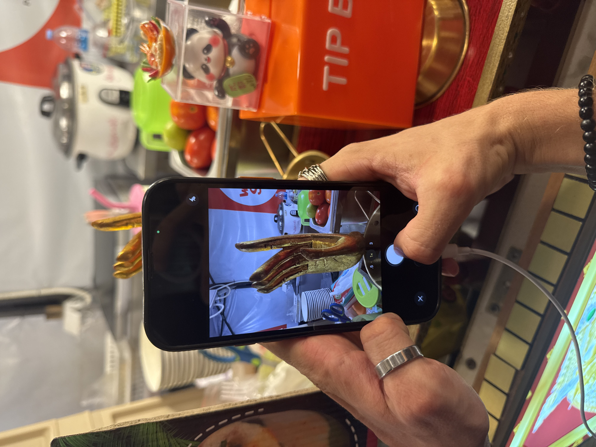 In Karon, Thailand, a person's hands are visible holding a smartphone, capturing an image of a golden hand-shaped sculpture. The sculpture is positioned in front of a collection of stacked white plates and various kitchen utensils, including blue and green spatulas. In the background, a white rice cooker and a green pitcher are present, along with a clear container holding a small panda figurine and a dish of food topped with shrimp. To the right, an orange tip box with embossed white lettering is prominent. The scene appears to be set in a busy market or food stall environment.