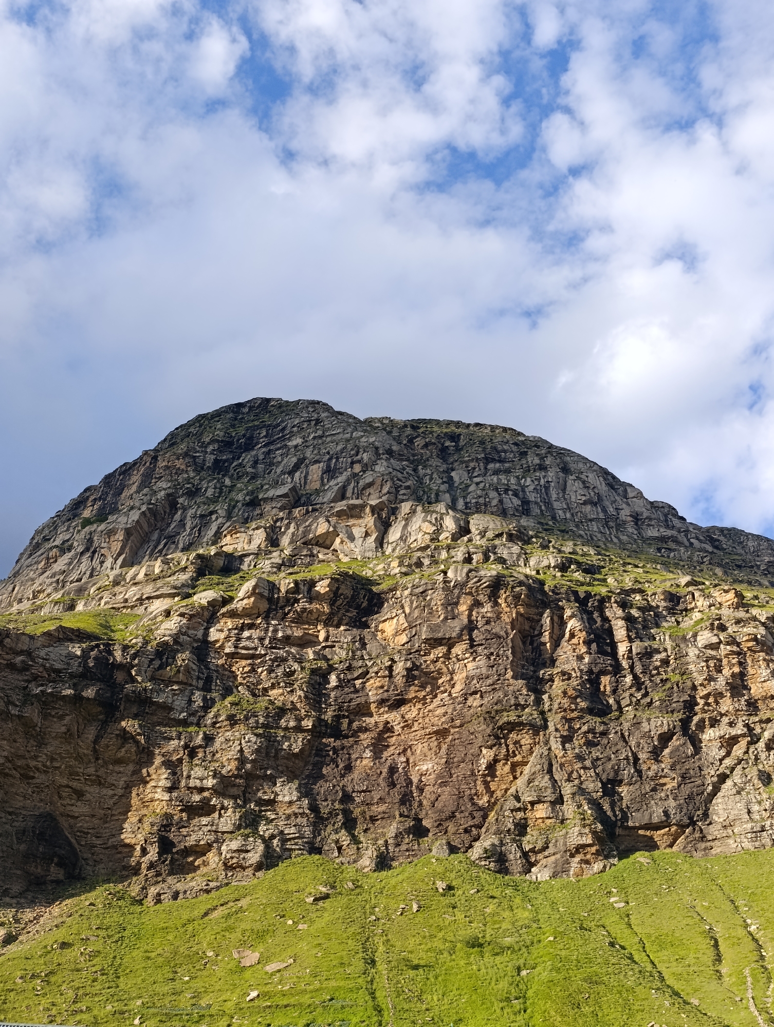 A low-angle, full shot captures a massive, rugged mountain with patches of green grass and vegetation clinging to its lower slopes. The mountain face is composed of layered rock formations, showing variations in color from dark gray and brown to lighter tan hues. Some sections appear sheer and imposing, while others are broken and irregular, with crevices and overhangs. Sunlight illuminates parts of the rock face, creating shadows that emphasize its texture and depth. The sky above is a bright blue, dotted with fluffy white clouds, suggesting fair weather. The location is identified as Damphug, India.