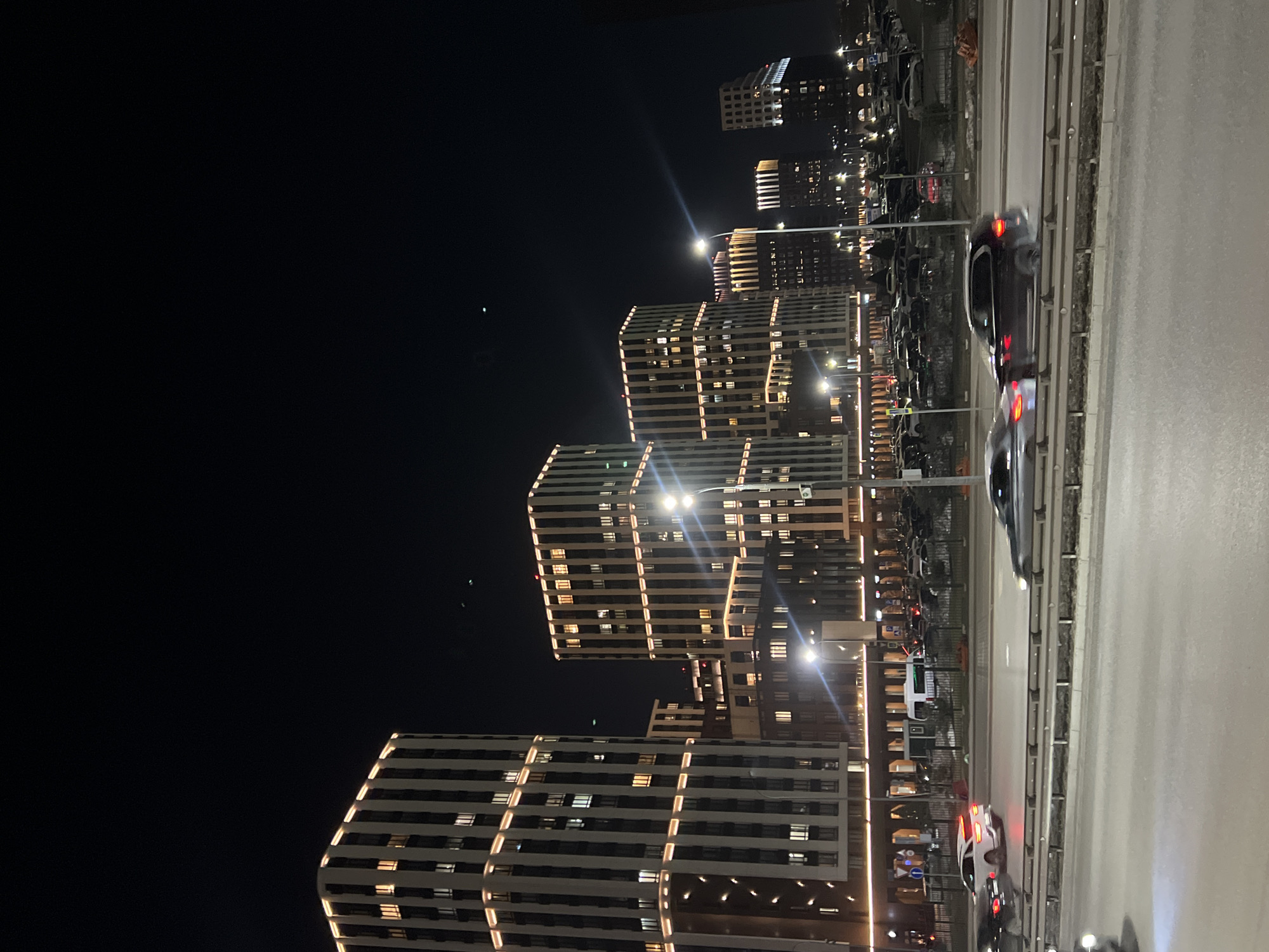 The image depicts a nighttime scene in Kazan, Russia, showcasing modern architecture alongside a highway. Several tall buildings are visible, characterized by their rectangular shape and horizontal lines of illuminated windows. Streetlights are positioned between the buildings and the road, casting a bright glow. The road has several cars driving on it. The night sky is black.