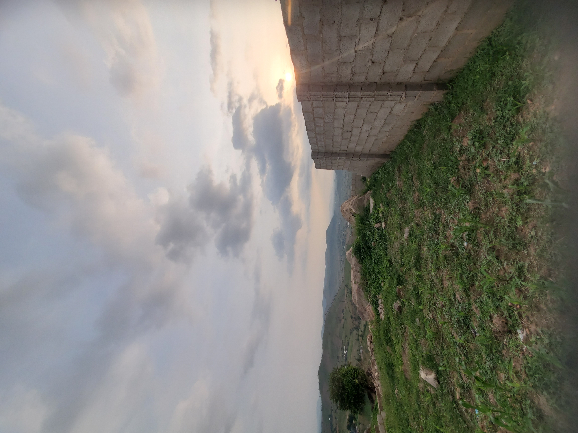The image captures a view from an elevated, undeveloped area overlooking the city of Suleja, Nigeria, at what appears to be sunset. In the foreground, a rough, unmortared brick wall runs diagonally from the lower right corner towards the middle of the frame, with sparse green vegetation growing around its base and on the uneven terrain to its left. The terrain slopes downwards to the left, revealing a landscape with rolling hills, scattered trees, and signs of development with some buildings visible in the distance. The sky is filled with dramatic clouds, with the sun setting on the right side, casting a warm glow and visible rays of light. The overall impression is one of a rural or semi-developed outskirts of Suleja, with natural elements juxtaposed against emerging construction. There are no people or animals visible in the scene.