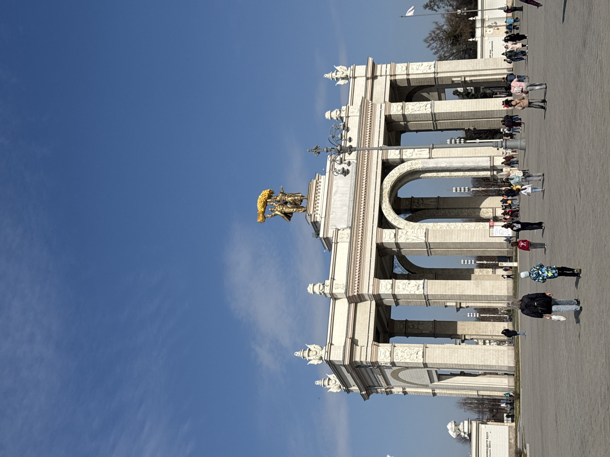 The image depicts an ornate, white-columned archway structure, possibly a triumphal arch, topped with a gilded sculpture of figures. This is most likely the main entrance to the Exhibition of Achievements of National Economy (VDNKh) in Moscow, Russia. The structure features multiple arches and decorative reliefs.
People are walking on the pavement to the right of the structure. They are dressed in casual clothing. A blue sky provides the backdrop.