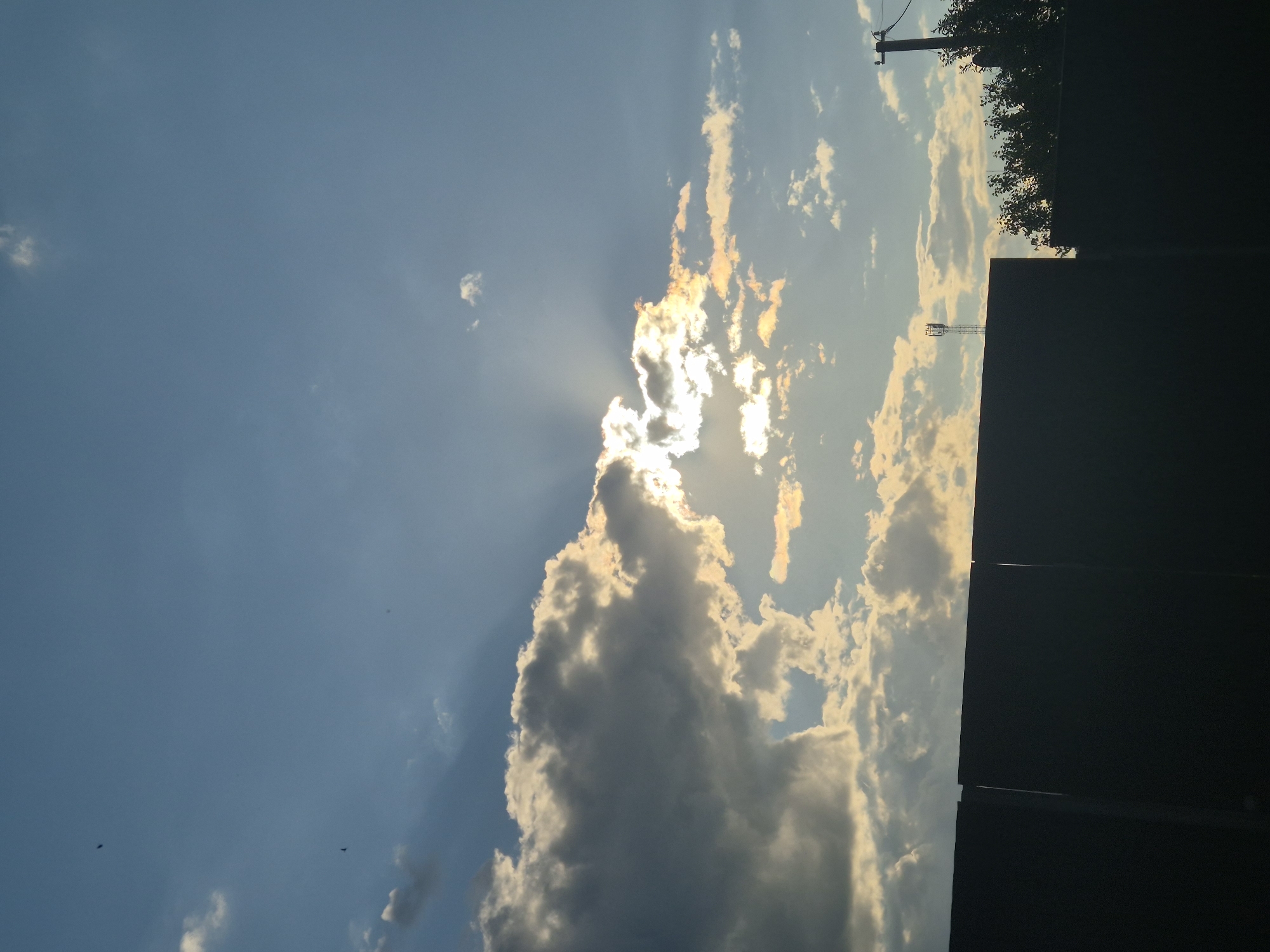 A wide shot captures a cloudy sky with the sun peeking through, casting light rays. In the foreground, a dark, silhouetted fence stretches horizontally across the bottom of the frame, with a gap in the middle. Behind the fence, a communication tower stands tall, and to its right, the silhouette of trees and a utility pole are visible. The sky transitions from a bright blue at the top to lighter tones near the horizon, with scattered clouds that are illuminated by the sun. The overall scene suggests an outdoor environment, possibly on the outskirts of a city, with elements of infrastructure and nature. The location is specified as Sozonivka, Ukraine.