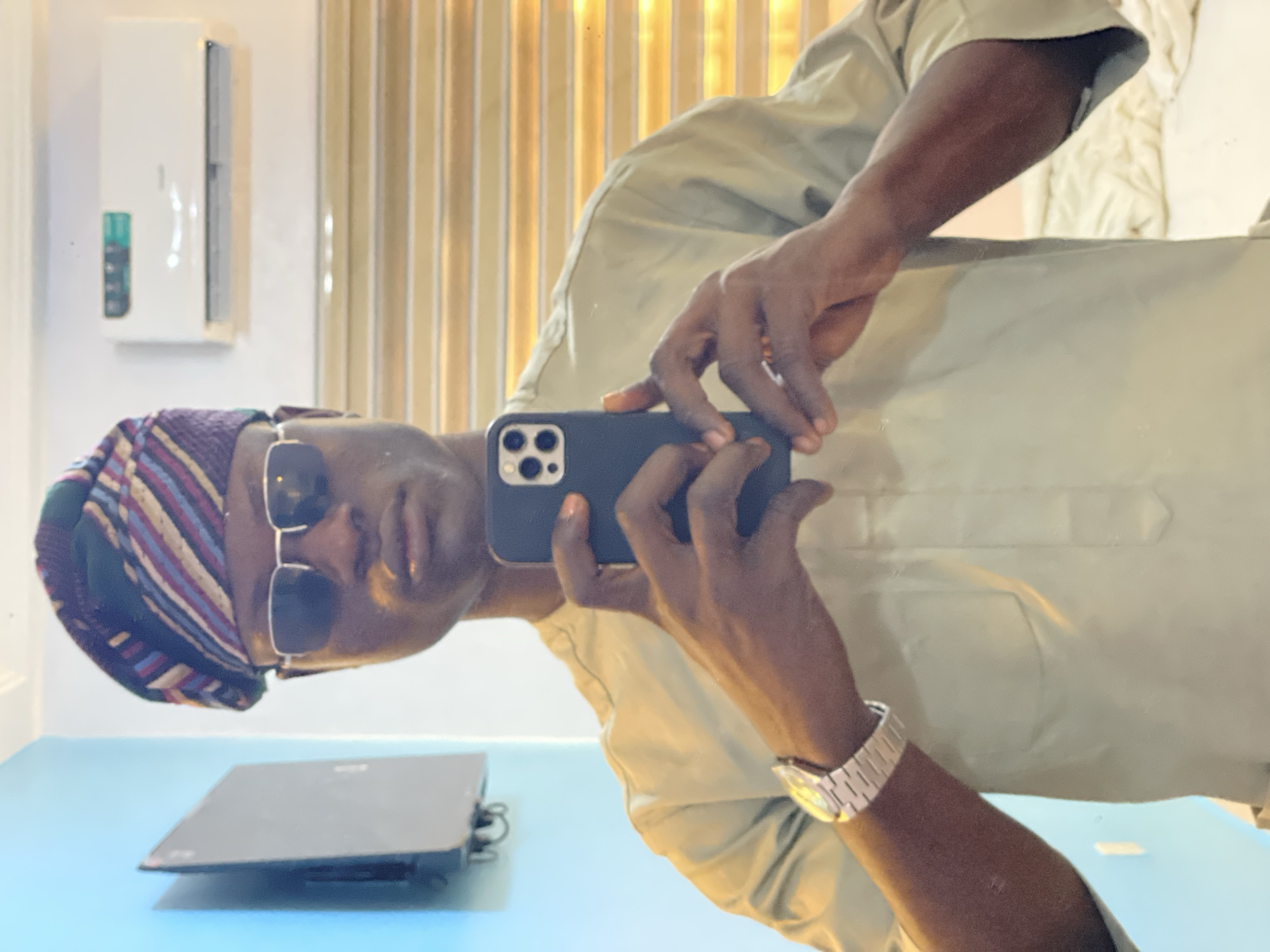 A person wearing a traditional Yoruba hat, sunglasses, and a light-colored shirt is taking a selfie with a smartphone. A watch is visible on their left wrist. The person is standing in front of a shiny surface, likely a mirror. A laptop sits on a turquoise surface in the foreground. In the background, vertical stripes are visible. A white box is mounted on the wall. The location is Badagry, Nigeria, but no specific landmarks are visible to provide more precise information.