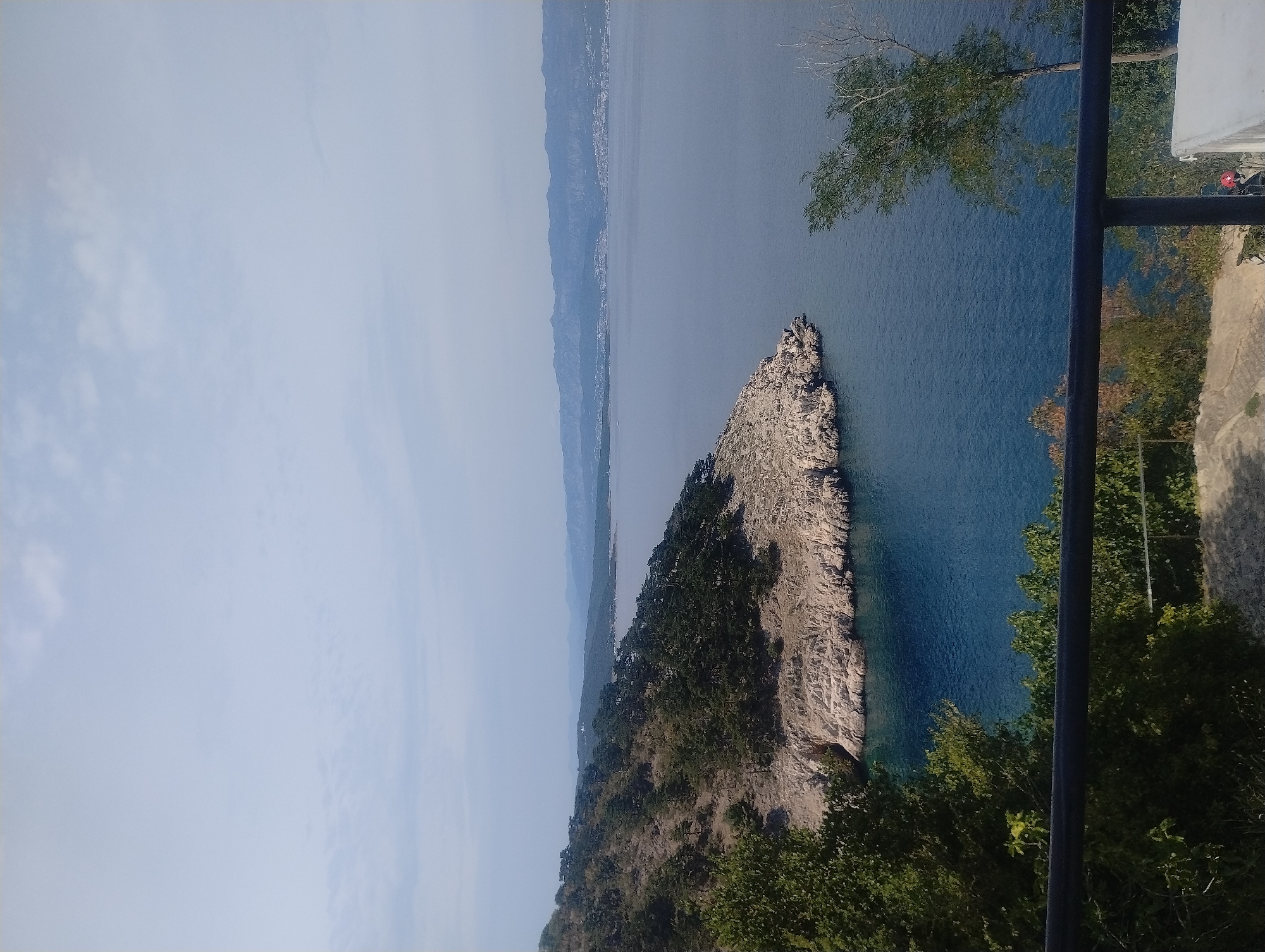 An elevated, wide-angle, eye-level, outdoor, daytime shot captures a scenic coastal view in Općina Vrbnik, Croatia. A rocky, tree-covered peninsula juts out into a calm, blue sea. In the distance, a hazy mountain range forms the horizon. In the foreground, a black metal railing runs horizontally across the bottom of the frame, partially obscuring lush green foliage and a stone path. A person wearing a red helmet is visible near the bottom right corner of the image. The sky is a pale blue with scattered clouds.