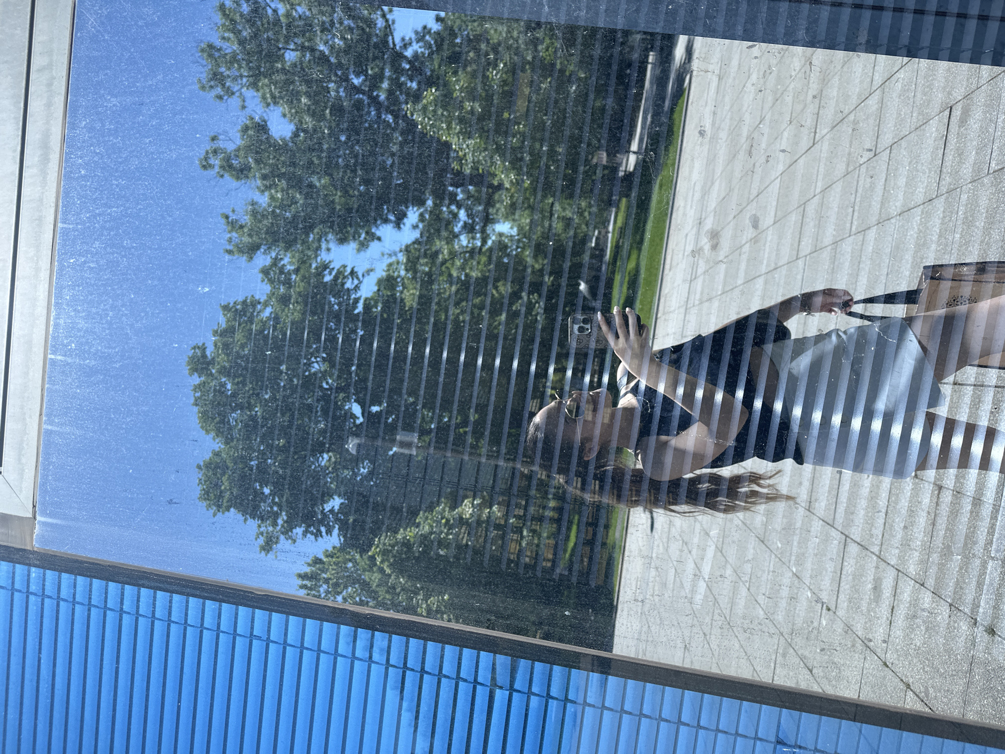 The image is a reflection in a window. The reflection shows a woman holding a mobile phone, presumably taking a photograph of herself. The woman is wearing a dark top and a light-colored skirt or shorts. Her hair is long and blonde.
The background reflected in the window shows trees and a paved area. The trees appear to be in a park or other green space. The sky is visible through the trees and is a clear blue.
The actual window appears to be part of a building with blue cladding. The blue cladding is ribbed or has a similar pattern.
It is impossible to confirm the location is Sofia, Bulgaria, without additional information or landmarks visible in the image.
