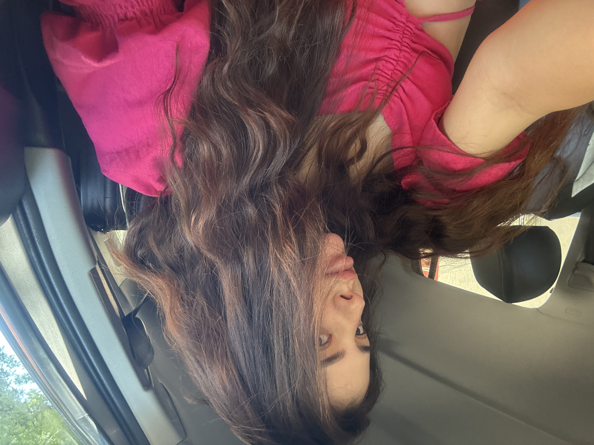 A young woman with long, dark, wavy hair, is seated in the passenger seat of a vehicle, looking to her left. She is wearing a bright pink, ruched top. The interior of the car is visible, with a headrest and door panel. Sunlight streams in from the right side, illuminating the scene. The image does not contain any information or visual cues that would allow for the identification of a specific location within the city of Plovdiv, Bulgaria.