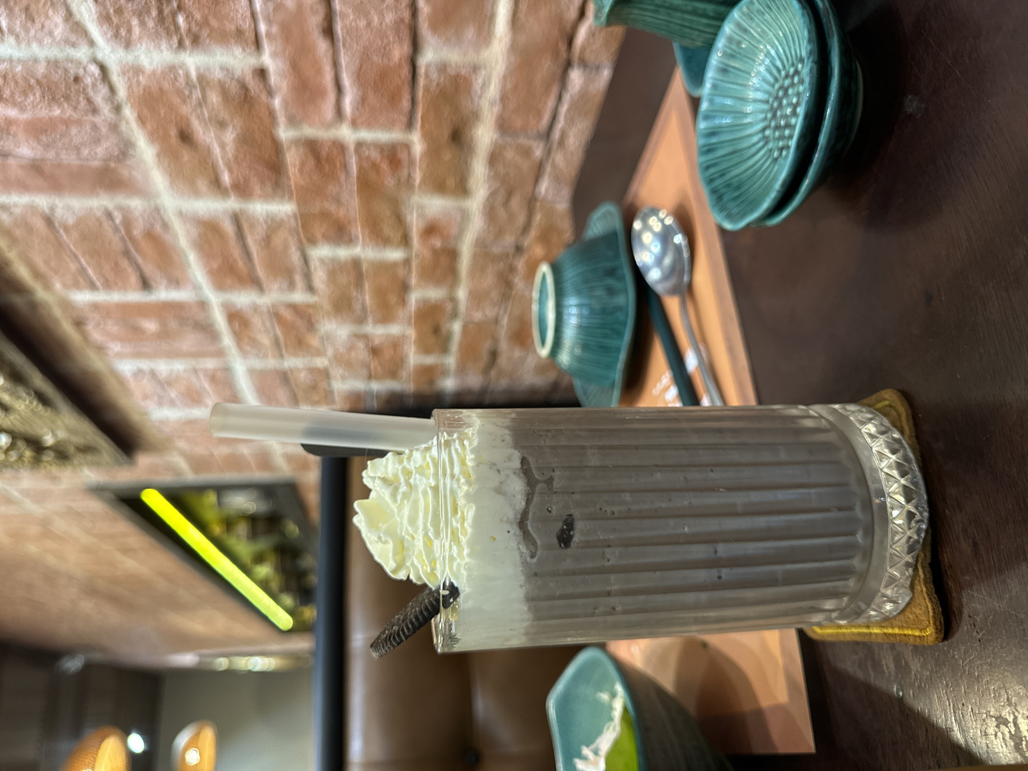 The image depicts a close-up shot of a chocolate milkshake topped with whipped cream and a cookie, served in a fluted glass on a coaster. The setting appears to be a restaurant, possibly in Phan Thiết, Vietnam, given the presence of what seems to be a traditional Vietnamese tea set with small green bowls and a spoon. In the background, a brick wall and a booth seat suggest a casual dining atmosphere. The lighting from overhead fixtures and a neon accent light creates a warm ambiance. Other elements in the scene include a glimpse of a salad in a teal bowl and a menu or placemat. The overall impression is one of a relaxed meal or snack in a restaurant.