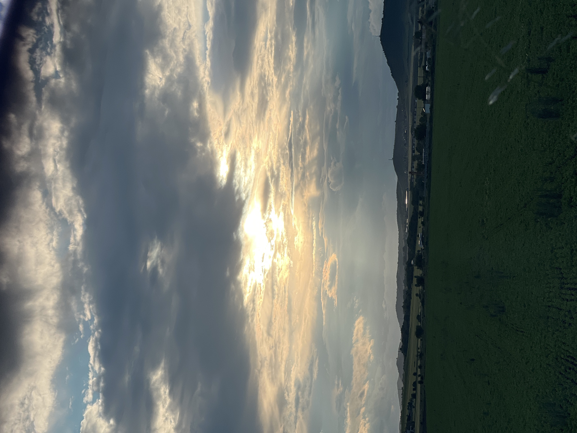 The image depicts a landscape with a bright, partly cloudy sky dominated by a dense cloud formation, revealing a strong sunlight source in the middle. The lower right of the image shows a green field, indicating likely agricultural land. In the background, a thin line of dark green vegetation is visible. Further into the horizon, traces of urban development can be observed, aligning with the geographical context of Aheloy, Bulgaria. No people are visible in the picture. The primary focus is on the interplay of natural elements: light, clouds, and landscape.