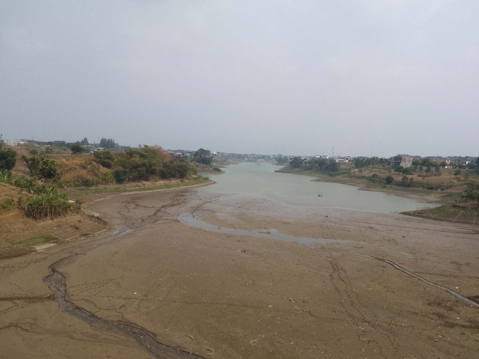 The image depicts a wide, muddy riverbed with a narrow stream of water flowing through the center. The banks of the river are dry and cracked, with sparse vegetation. In the background, there are houses and trees on a hill. The sky is overcast. The scene is consistent with the dry season in Bojonghaleuang, Indonesia, where water levels in rivers and reservoirs often recede, exposing large areas of mud. The presence of houses on the hillside suggests a residential area near the river.
