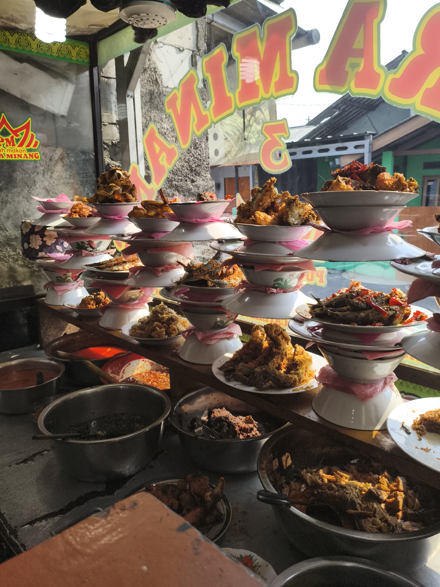 The image depicts the interior of a small, casual Indonesian restaurant, likely a Nasi Padang establishment, in Depok, Indonesia. A prominent sign with red and yellow lettering, partially obscured by reflection, reads "Nasi..." and likely continues with "Minang" or a similar identifier, suggesting it is a restaurant specializing in Minangkabau cuisine.
The foreground shows several large metal bowls filled with various dishes. One bowl on the left contains a reddish sauce or soup. Adjacent to it is a large, dark bowl filled with a dark, stew-like food, possibly beef rendang or a similar slow-cooked meat dish, with a metal spoon resting inside. Another bowl to the right holds a dark, chunky mixture, and further to the right, a white plate with a portion of a dish is visible.
The middle ground features stacked plates and bowls, a common display method in Nasi Padang restaurants, showcasing an assortment of prepared dishes. These include fried chicken, fish, and various curried or sauced items, presented in white bowls and plates, often with pink paper liners underneath. The dishes are arranged in tiered stacks, creating a visually abundant display.
The background shows the exterior environment through a glass window. Sunlight streams in, creating reflections on the glass. The outside scene includes glimpses of buildings with corrugated metal roofs, suggesting a typical urban or suburban Indonesian setting in Depok. A smaller logo on the left side of the window, featuring red and green elements, further identifies the establishment. The overall impression is one of a bustling, locally run eatery offering a wide variety of traditional Indonesian dishes. No people are directly visible in the image.