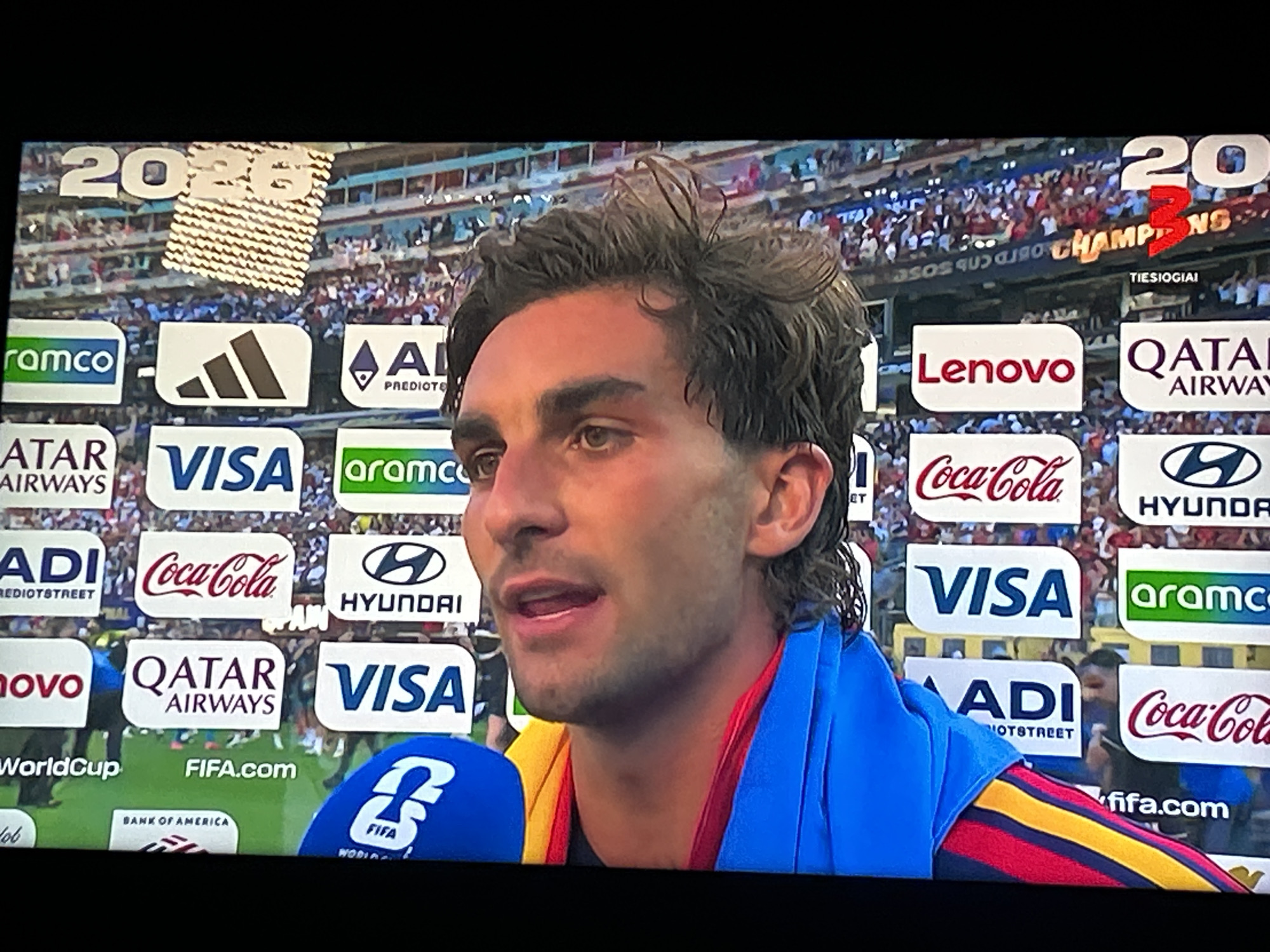 A medium shot captures a man from the chest up, speaking into a microphone. He has brown, wavy hair and is looking to his left. He is wearing a blue towel or scarf draped over his shoulders, and a multi-colored striped shirt or jacket underneath. Behind him, a stadium crowd is visible, with numerous sponsor logos on display. These logos include "FIFA.com", "VISA", "aramco", "Coca-Cola", "Lenovo", "Qatar Airways", "Adidas", and "Hyundai". The backdrop also features a large "2026" sign and a "3 CHAMPIONS" sign. The overall setting appears to be a sports event, likely a soccer match, in a large stadium.
