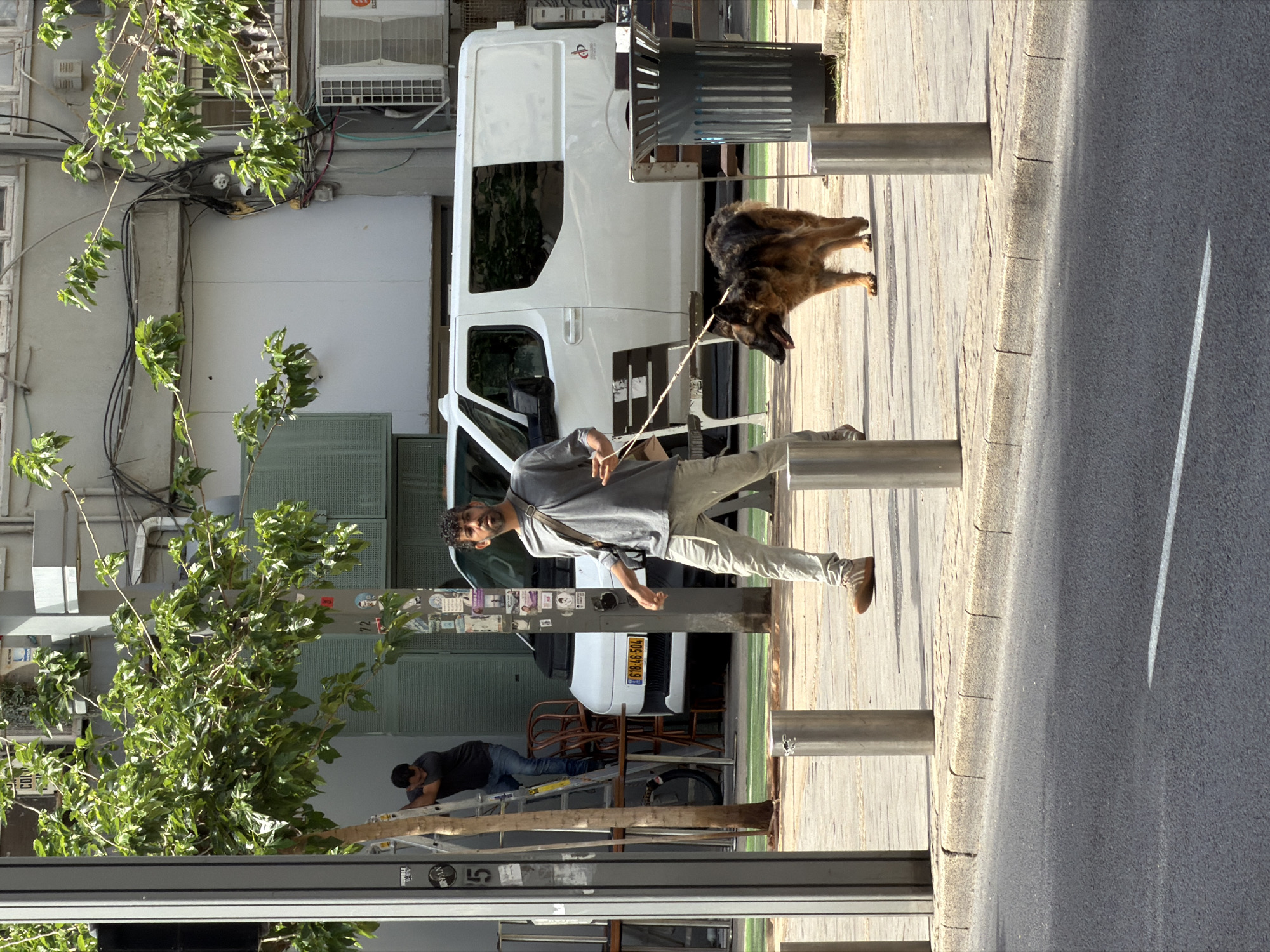 A man walks his dog on a Tel-Aviv sidewalk next to a street. The dog is a German Shepherd. The man is holding a leash and wearing a t-shirt and pants. Another man is seen working on a ladder next to a van. There are trees and buildings in the background. A white line is visible in the lower right of the picture.