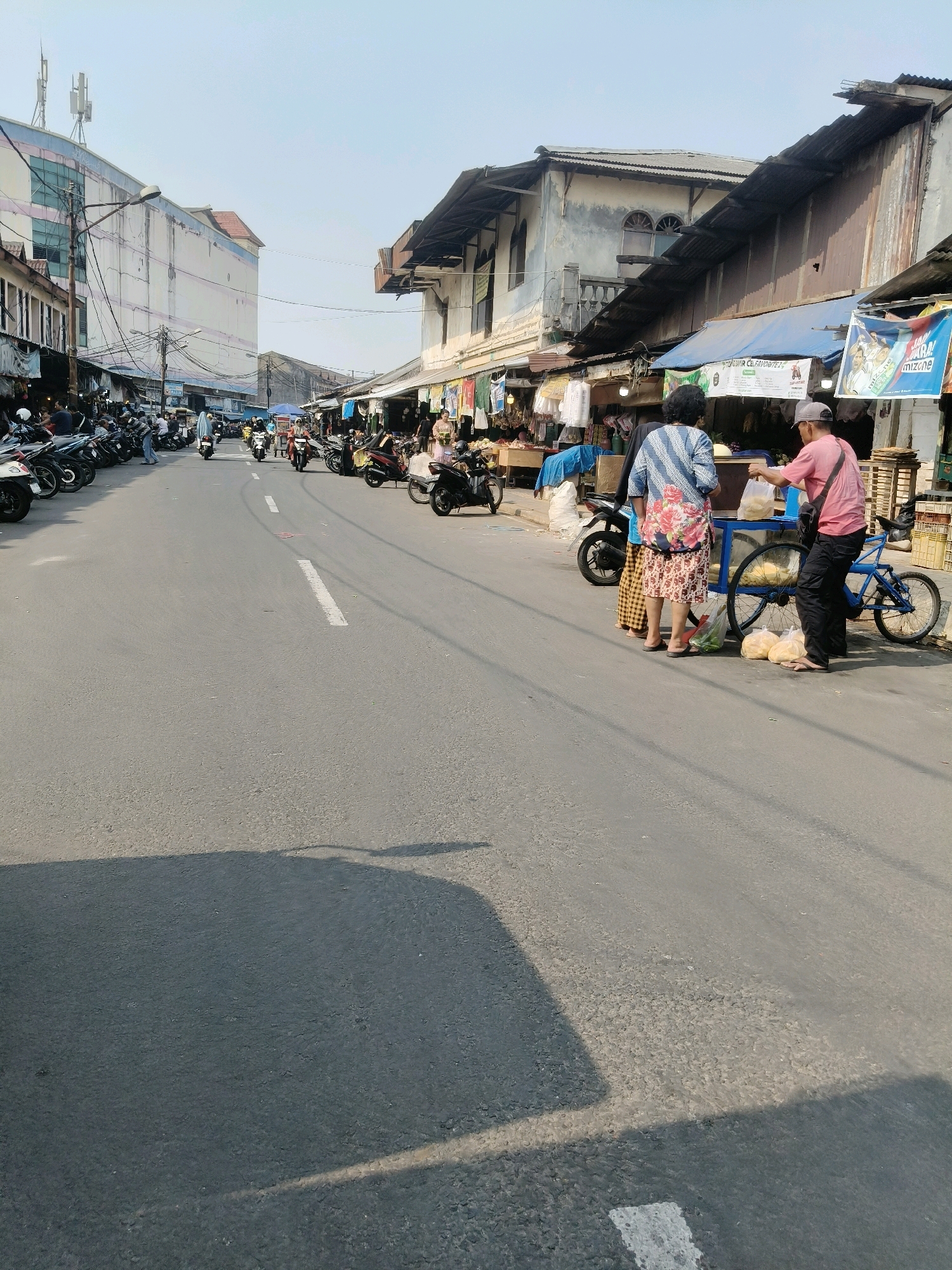 This image captures a bustling street scene in South Tangerang, Indonesia, likely a market area. The road, a typical asphalt surface with white lane markings, is flanked by numerous parked motorcycles on the left and a row of shops or market stalls on the right. The stalls have corrugated metal roofs and are adorned with various goods, including clothing and produce, suggesting a commercial environment.
In the foreground, two individuals are engaged in an interaction. A person wearing a pink shirt and a baseball cap is handing something, possibly food items in plastic bags, to another individual wearing a patterned blue and white shirt and a floral skirt. They stand near a cart with two bicycle wheels, which appears to be a vendor's cart. Several plastic bags filled with what seem to be snacks or produce are also visible on the ground near the cart.
Further down the street, a steady flow of motorcycles navigates the road, indicating active local transportation. The buildings in the background vary in style, with some appearing modern with multiple stories and antennae on the roof, while others are older, characterized by weathered facades and traditional architecture. The sky is clear and bright, suggesting it is daytime. The overall atmosphere is one of everyday commerce and activity within a developing urban setting.