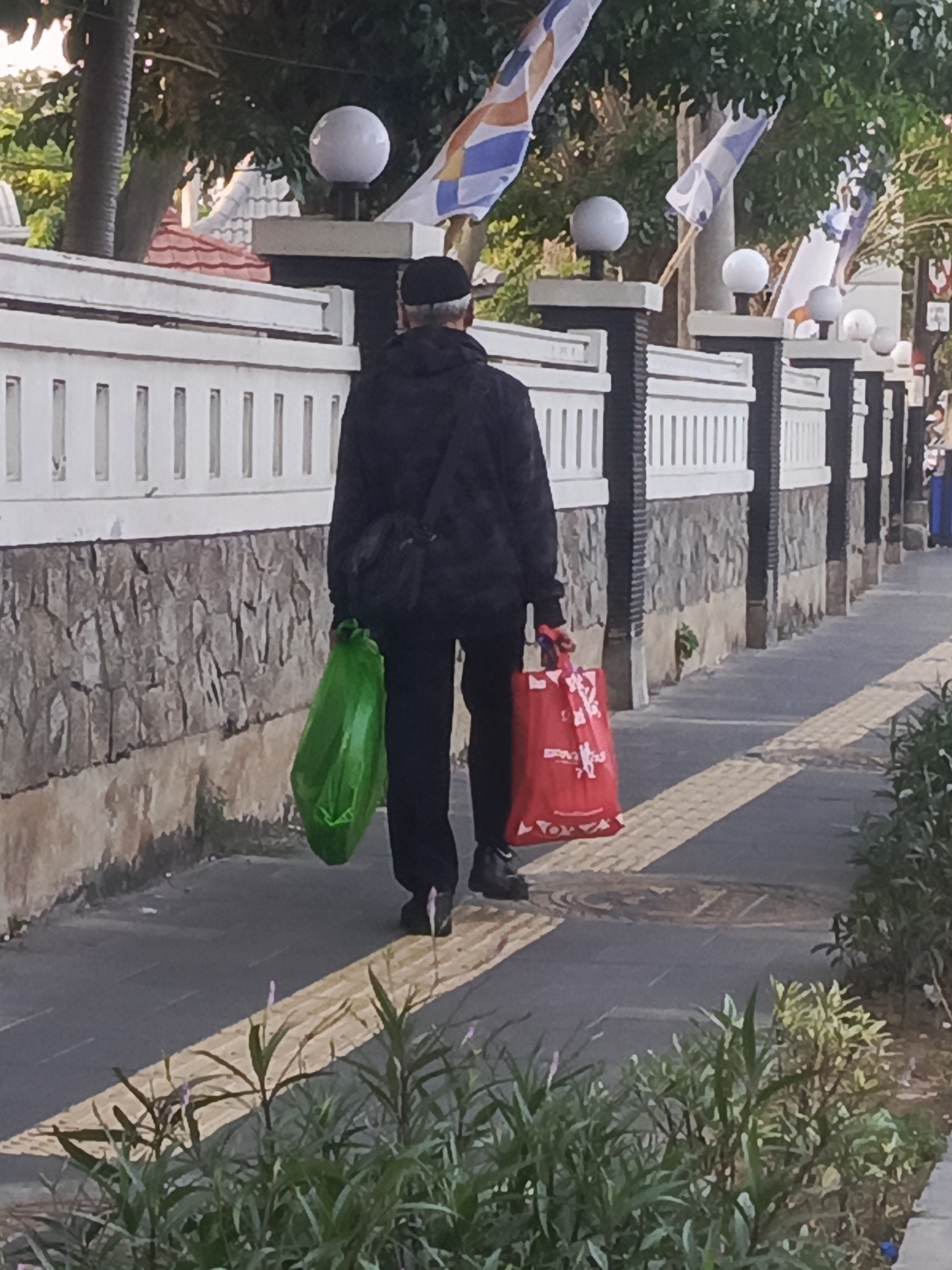 A person walks along a sidewalk in Surabaya, Indonesia, carrying two shopping bags. The person is wearing a black hooded jacket, black pants, and a black cap. They are walking away from the camera, and their face is not visible. One bag is green and the other is red. The sidewalk is paved and has a yellow tactile paving strip. Behind the person is a stone wall and a white fence. There are flags and lights visible on top of the fence. The area is outdoors, and it appears to be daytime. Lush green plants are growing along the edge of the sidewalk.