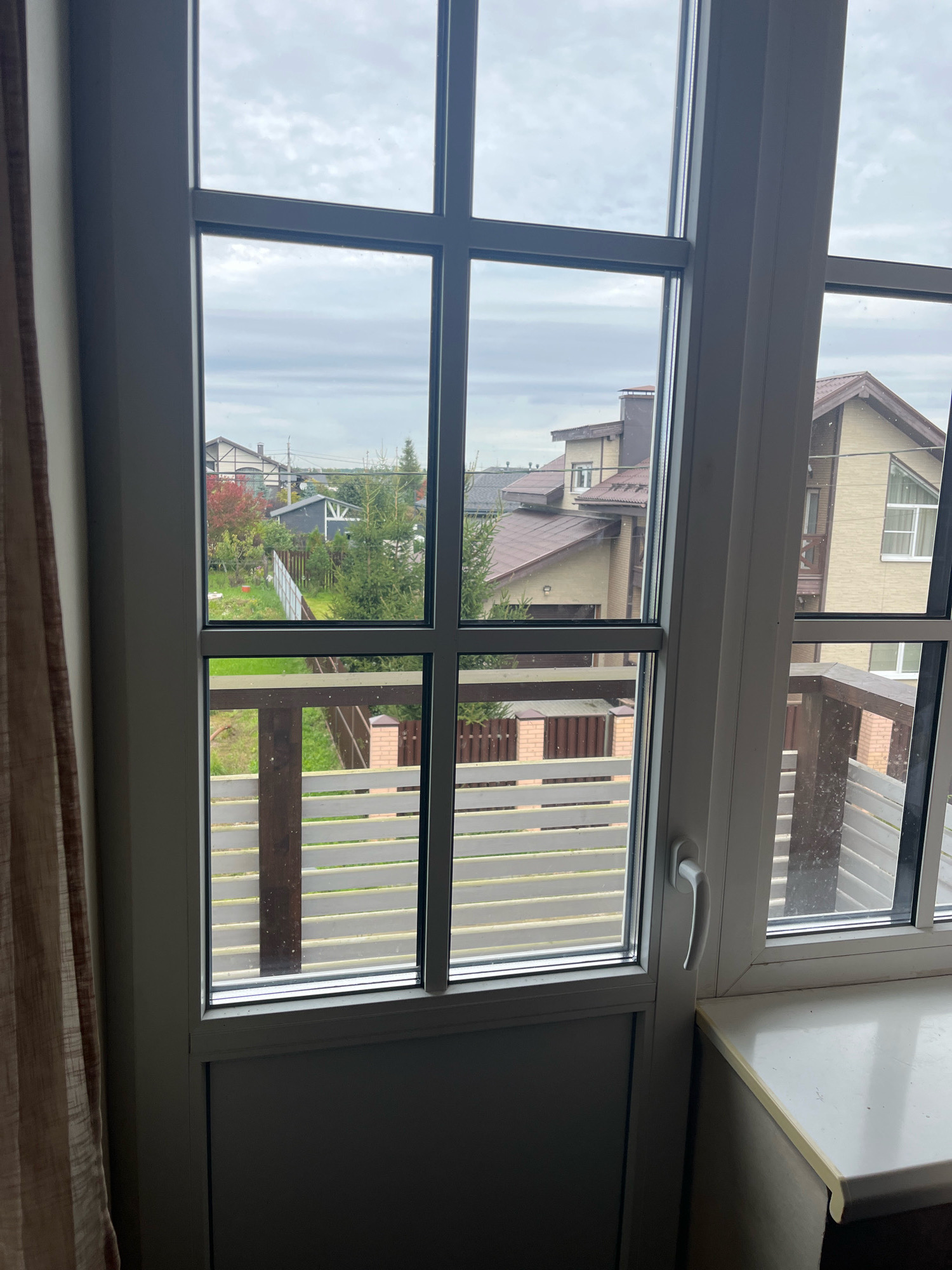 This is a medium shot, eye-level, indoor exterior shot of a window overlooking a residential area. The window is large, with multiple panes, and is framed in white. The view outside shows a cluster of houses with reddish-brown tiled roofs. There are some evergreen trees and deciduous trees with autumn foliage, suggesting the season is fall. A wooden fence runs along the property line. A white railing with wooden posts is visible on a balcony or porch. The sky is overcast with gray clouds. The bottom part of the window reveals a darker interior, possibly a room or hallway, with a light-colored windowsill. A beige curtain is partially visible on the left side of the frame. The scene appears to be in a suburban or rural setting, possibly a dacha or country house area. The mention of Vladimirovka, Russia, would place this scene in that specific geographic context.