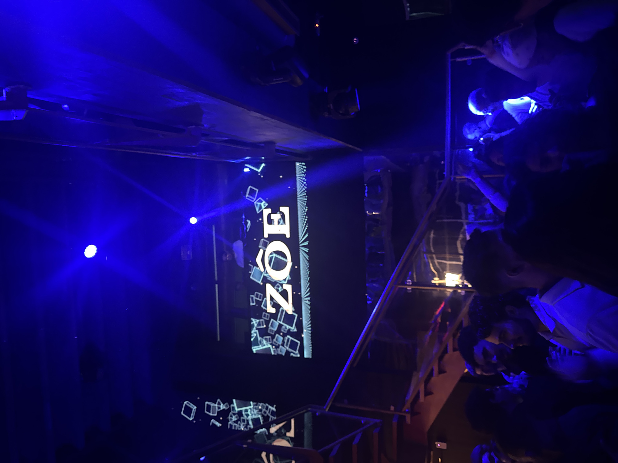 The image shows a crowded indoor setting, possibly a nightclub or bar in Caracas, Venezuela. The lighting is dominated by blue hues, with spotlights illuminating various areas. A large vertical screen displays the word "ZOE" in white capital letters, surrounded by geometric shapes that appear to be 3D cubes. A staircase is visible on the right, with people standing on it, some interacting with each other. The overall atmosphere appears to be lively and energetic.