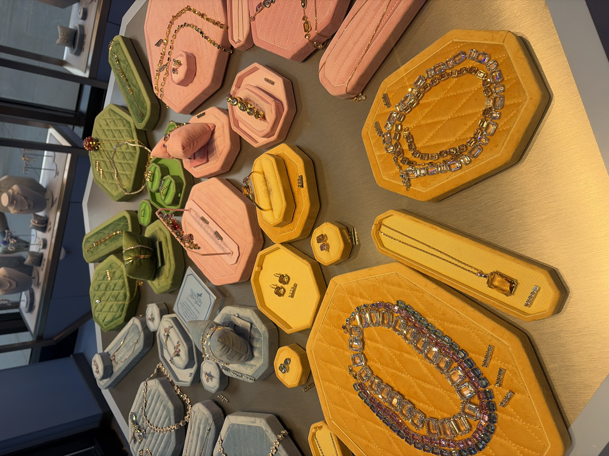The image shows a display of jewelry, likely in a retail environment, with various necklaces, bracelets, and earrings presented on plush velvet trays in shades of blue, green, pink, and yellow. The jewelry features numerous gemstones, many of which appear to be faceted and possibly diamond or crystal. Some pieces are intricate and multi-layered. The background reveals glass display cases with more jewelry items and mannequins, suggesting a high-end jewelry store. The lighting is bright and focused on the merchandise, creating a polished and luxurious presentation. The specific location in Montreal, Canada, cannot be determined from the visual information provided. There are no people or actions depicted in the image, only static displays of jewelry.