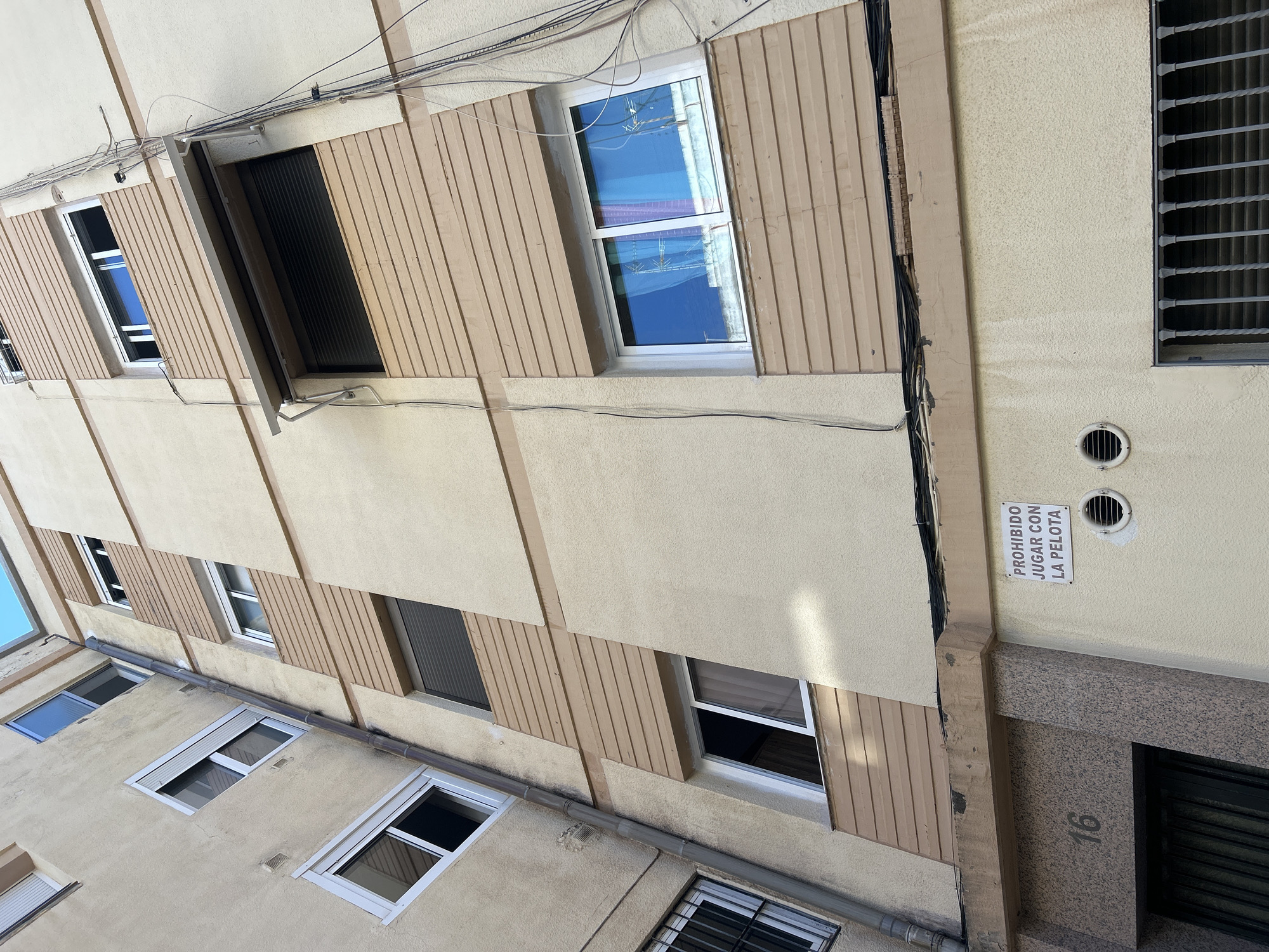 The image shows an exterior view of an apartment building, likely in Castelló De La Plana, Spain, as indicated by the context. The building facade is a light beige color with decorative wooden panels in a lighter tan, arranged vertically and diagonally. There are numerous windows visible, some open and some closed, with white frames. Some windows have shutters, and one on the upper right has a decorative metal grate. Electrical wires are strung across the top of the building, and a drainage pipe runs horizontally across a section of the facade.
One notable detail is a white sign attached to the wall near the entrance of the building. The sign has black text that reads "PROHIBIDO JUGAR CON LA PELOTA," which translates from Spanish to "PROHIBITED TO PLAY WITH THE BALL." This suggests a rule or warning related to activities in the area. A circular vent is visible above and to the right of the sign. The entrance area features a granite-like textured material, and the number "16" is stenciled on this surface.
The lighting suggests it is daytime, with shadows visible on the building, indicating sunlight. The sky, visible through an open window, appears blue. The scene depicts a residential building, likely in an urban or suburban setting. No people are visible in the frame, and the overall impression is one of a typical building facade on a clear day.
