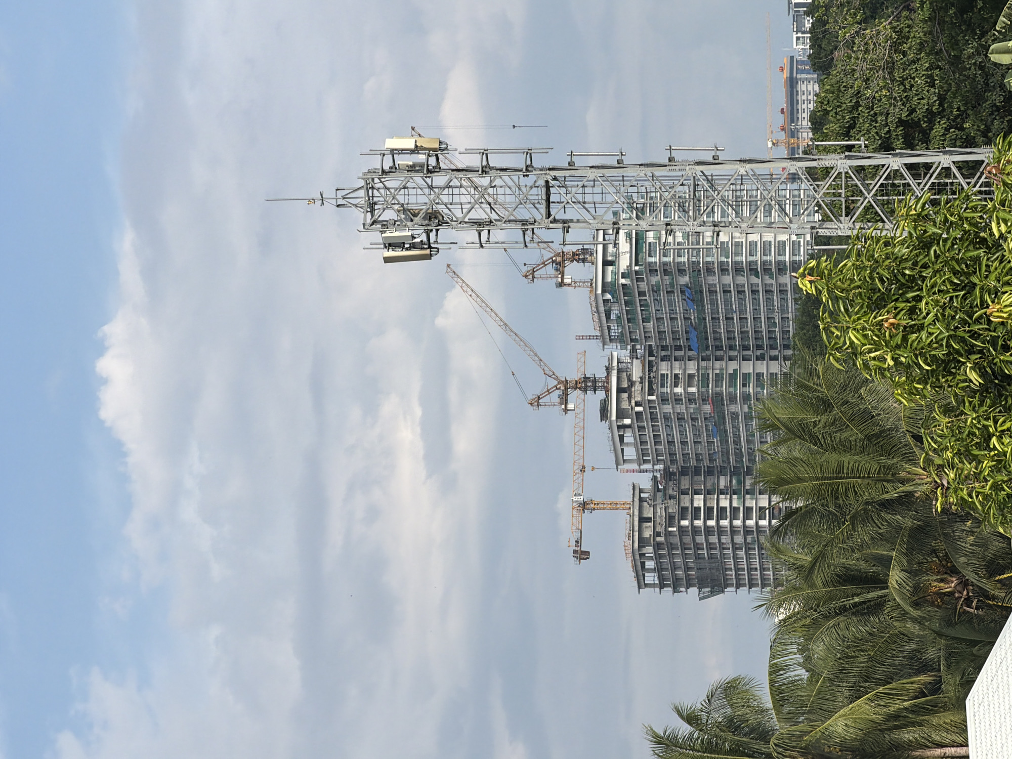 The scene features a construction site in Davao City, Philippines. A tall building is under construction, with exposed concrete structure and numerous floors visible. Several yellow construction cranes are positioned around the building. A lattice-like radio tower stands prominently in the foreground. Lush green foliage, including palm trees and other tropical vegetation, frames the lower and right portions of the image. The sky is a mix of light blue and cloudy patches.
