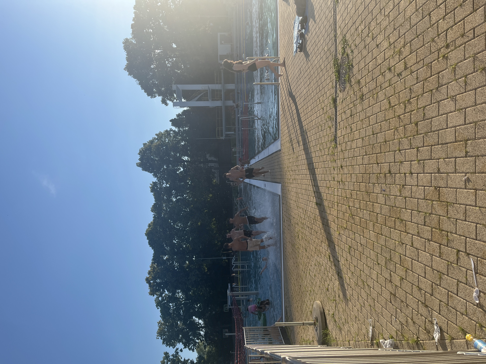A group of young people are running into a swimming pool under a clear blue sky. The pool has multiple lanes, a diving board structure, and is surrounded by a tiled patio. Trees line the perimeter of the pool area. The sun is setting, casting long shadows across the patio. Some trash is visible on the ground near the fence.