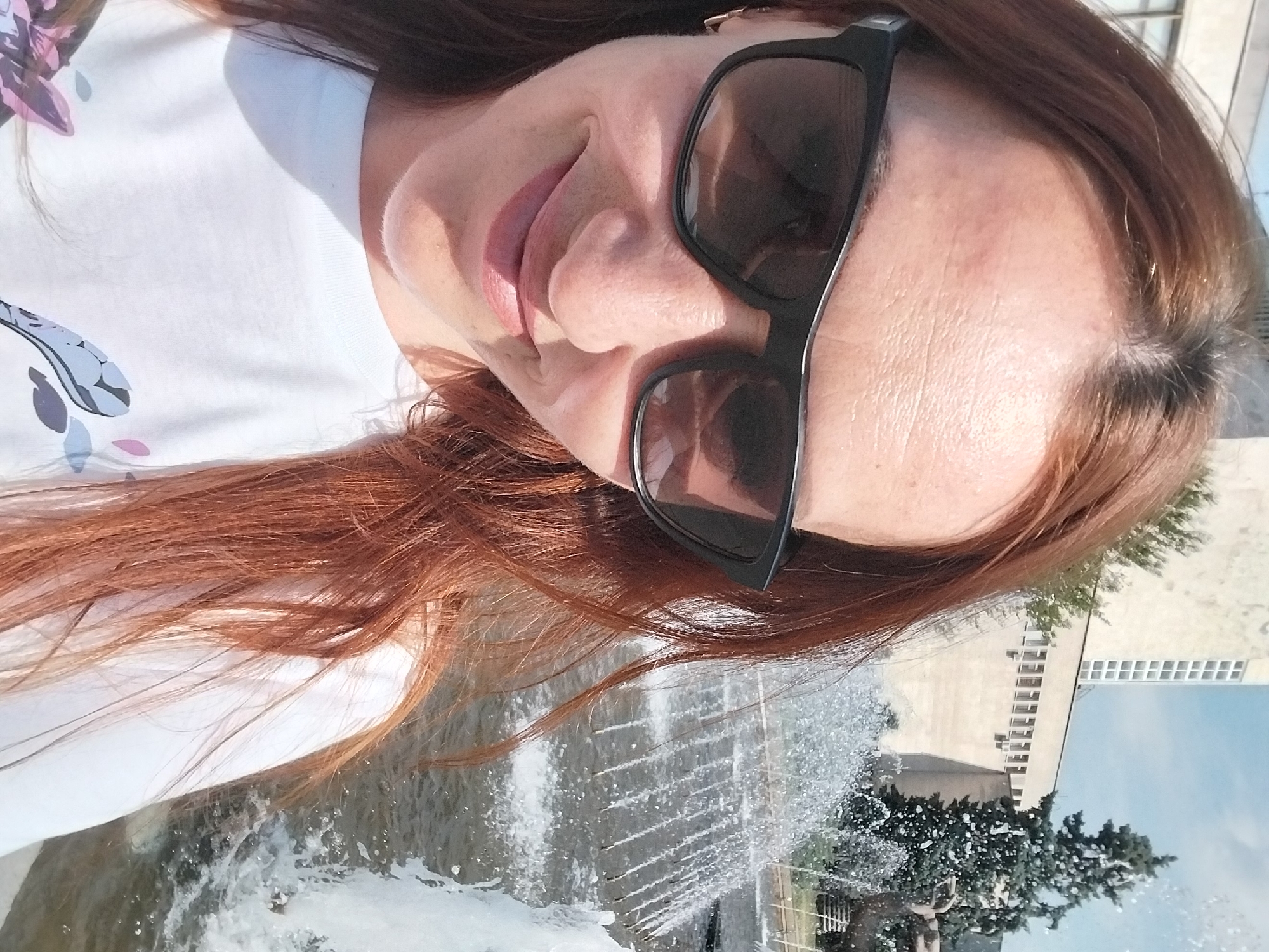 A woman with red hair, wearing sunglasses and a white t-shirt, stands in front of a fountain. The fountain is spraying water upwards, creating a mist. In the background, there is a building and a tree. The scene appears to be in a city park. This image is likely taken in the city of Dnipro, Ukraine.