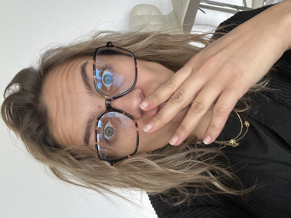 An adult individual, identified as female, is depicted in a close-up indoor portrait taken in Veauchette, France. Her light brown hair is disheveled. She wears large, square, tortoiseshell-patterned eyeglasses, with prominent reflections of a light source visible in both lenses. Her eyes are wide open, directed forward; the left iris appears greenish-blue, and the right appears blue. Horizontal creases are visible across her forehead. Her right hand, featuring natural nails with some wear and a small red mark near the index finger cuticle, is positioned over her mouth, covering her lips and lower nose. She is wearing a black top and a layered gold necklace with multiple small flower-shaped charms. The background consists of a plain white wall, with the upper portion of a white high-backed stool or chair visible on the far right.