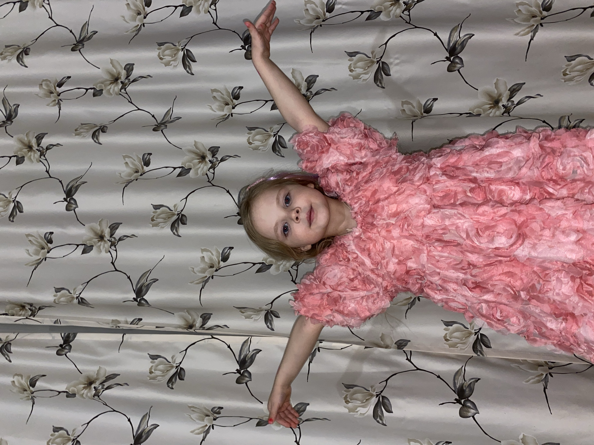 A young child is lying on a bedspread with a cream-colored background and a pattern of large white flowers with grey leaves and black stems. The child is wearing a pink dress with ruffled fabric. The child is fair-skinned with blonde hair and blue eyes. Her right arm is extended upwards and her left arm is extended downwards. The backdrop indicates an indoor setting. No information in the image indicates it is located in Votkinsk, Russia.