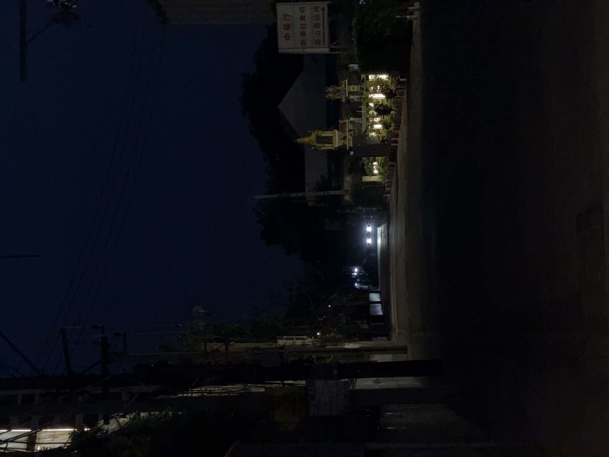 At night in Bangkok, Thailand, a street view shows illuminated shrines and potted plants lining the street next to a dark building. Power lines and utility poles are visible against the dark blue sky. Vehicle headlights illuminate the road further down the street. A sign with Thai script is mounted on a building.