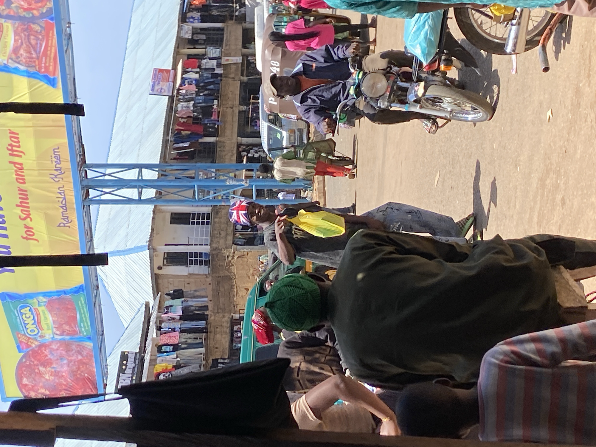 Captured in the city of Bukuru, Nigeria, the image shows a bustling street scene on a sunny day. People, including a man on a motorcycle, are present, along with various objects such as buildings, clotheslines with garments hanging, and advertising banners. An "ONGA" advert and a Ramadan greeting are visible. The scene features a vibrant array of colors and textures.
