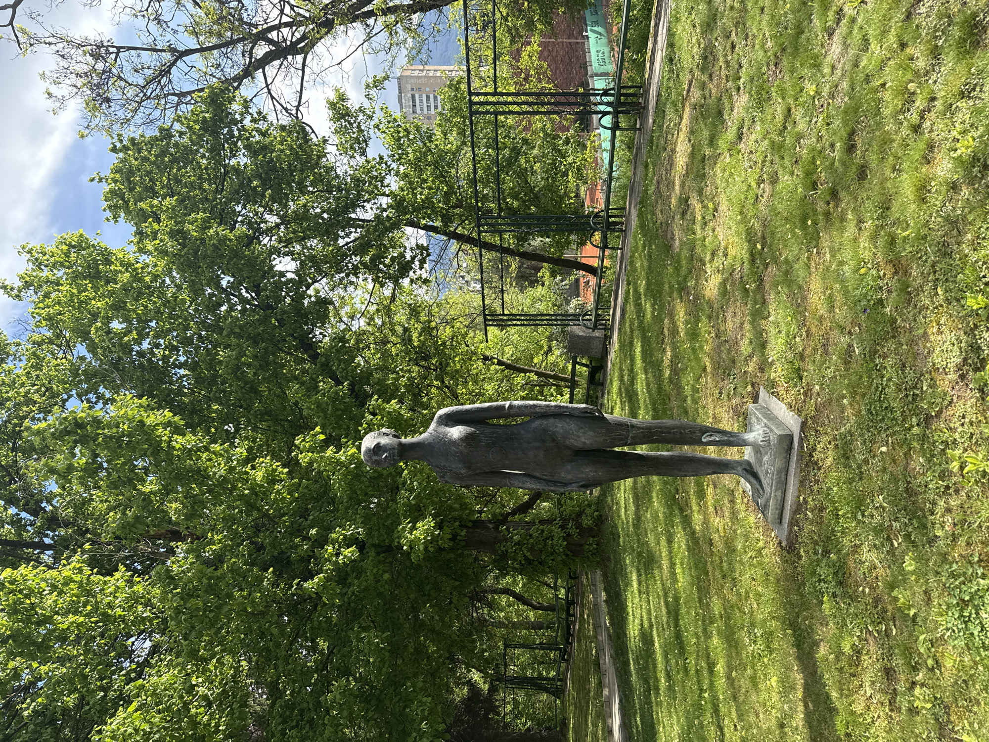 The image shows a bronze sculpture of a female figure standing in a park in Ljubljana, Slovenia. The sculpture stands on a stone plinth on a grassy area, with mature trees and a metal pergola structure in the background. A building is visible in the distance behind the trees and pergola. The weather appears sunny and bright.