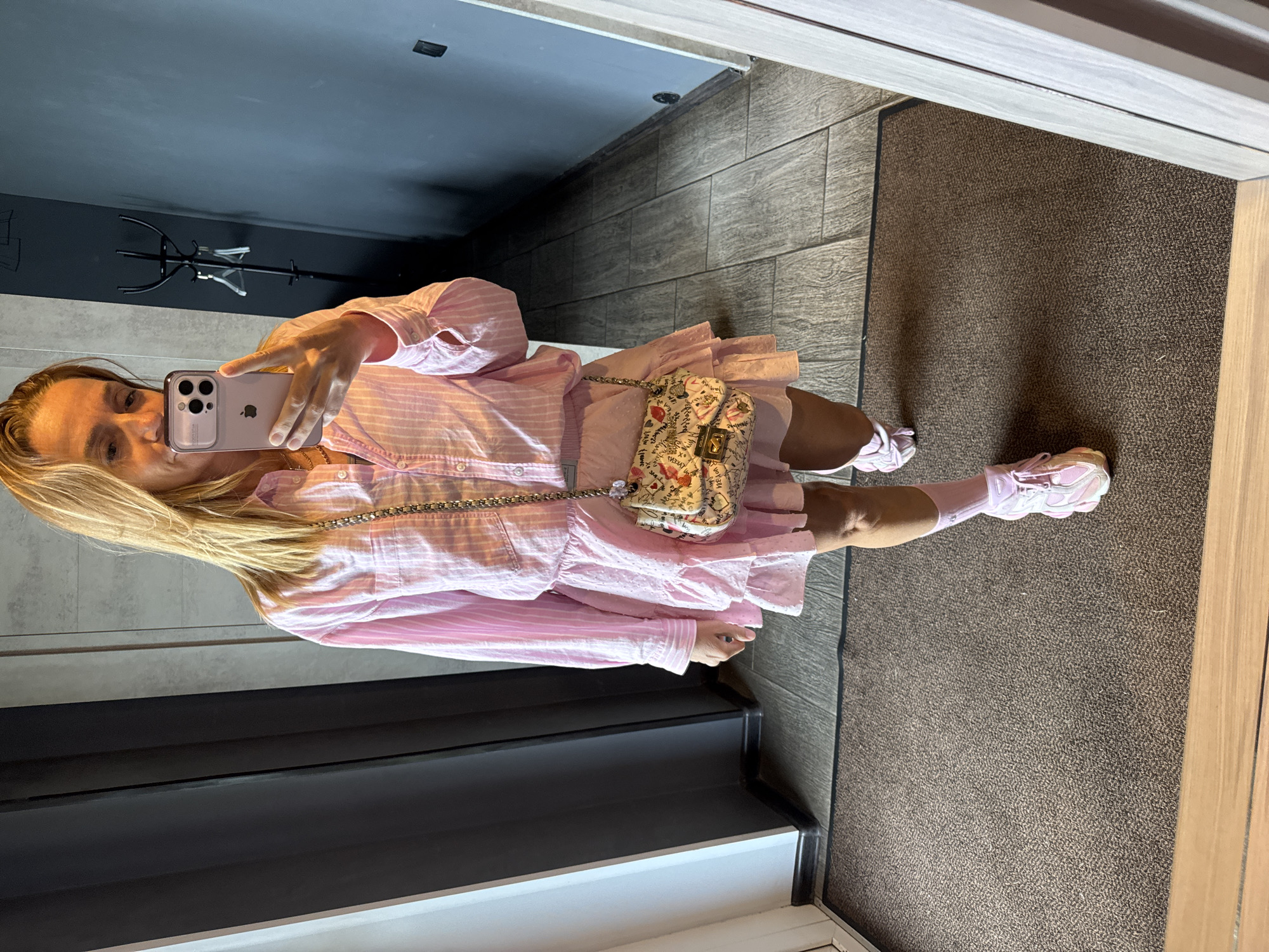 A blonde woman stands in a hallway, taking a selfie in a mirror. She is wearing a pink striped button-down shirt and a matching pink ruffled skirt. A small, white handbag with a floral and text pattern hangs from her shoulder. She has on pink socks and chunky pink and white sneakers. The hallway has dark grey walls, a tiled floor, and a black coat rack with hangers. There is a brown carpet on the floor in the foreground. The woman is looking at her phone, which she is holding up to take the picture. The environment suggests an indoor, possibly residential or hotel setting. There is no visible indication of the location being in Kirishi, Russia.