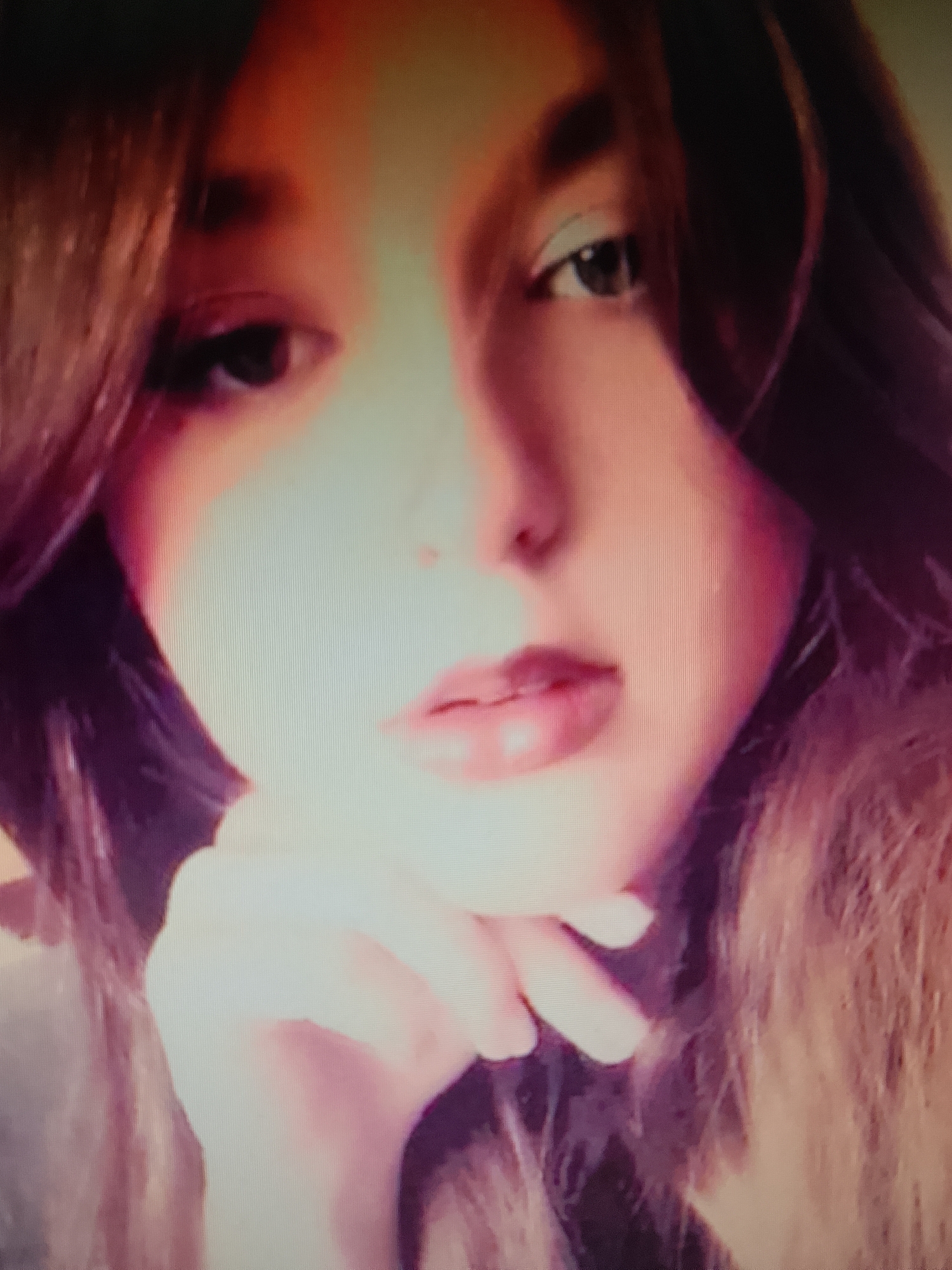 The image is a close-up, pixelated, and heavily filtered portrait of a young person. The subject's face is partially illuminated, with a strong light source from the front, creating bright highlights and deep shadows. The person has dark hair that falls around their face. Their right eye is visible, with dark irises and what appears to be light-colored eyeshadow. The left eye is mostly obscured by hair. The person's lips are slightly parted and appear to be painted with a glossy lipstick. Their right hand is raised, with the fingers resting on their chin, supporting their head. The background is indistinct and appears to be a soft, light color, possibly a wall or fabric. There are no recognizable landmarks, objects, or environmental features that would allow for specific identification of a location within Ostróda, Poland. The overall impression is of a selfie or a posed photograph with strong digital manipulation.