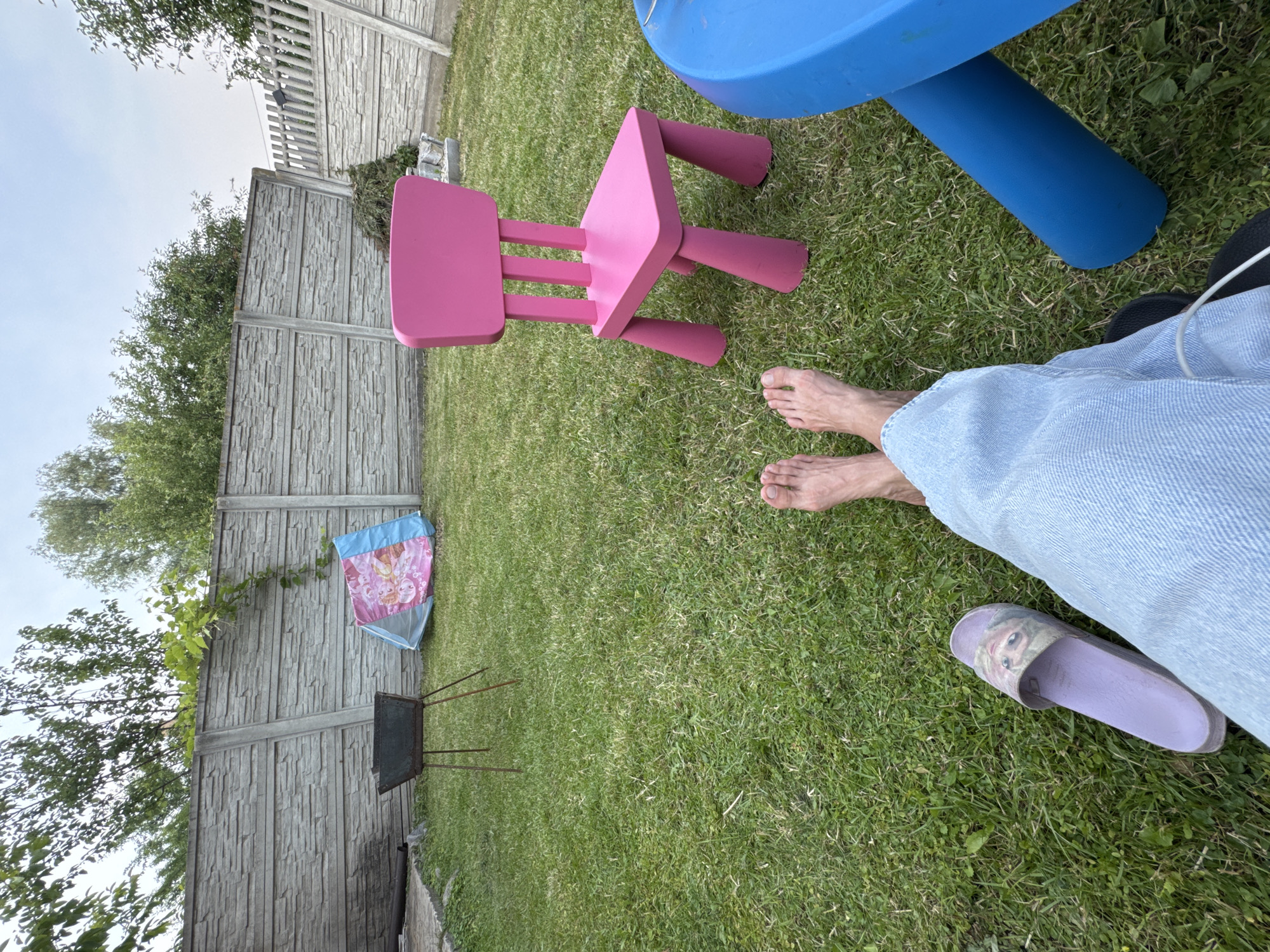 The image depicts a backyard scene with a person's legs and feet visible in the foreground, wearing light blue jeans and a purple slide with a cartoon character printed on it. To the right, a bright blue table leg is partially visible. In the middle ground, a pink child-sized chair with a rectangular back is prominently placed on the green grass. Behind the chair, a small tent with a pink and blue design, possibly depicting cartoon characters, is set up. To the left of the tent is a dark metal barbecue grill on three legs. A grey concrete fence forms the backdrop of the yard, with some greenery and trees growing over it and beyond. The sky is overcast. The scene appears to be a casual outdoor setting, possibly a residential backyard in a suburban or rural area, rather than a specific city location.
