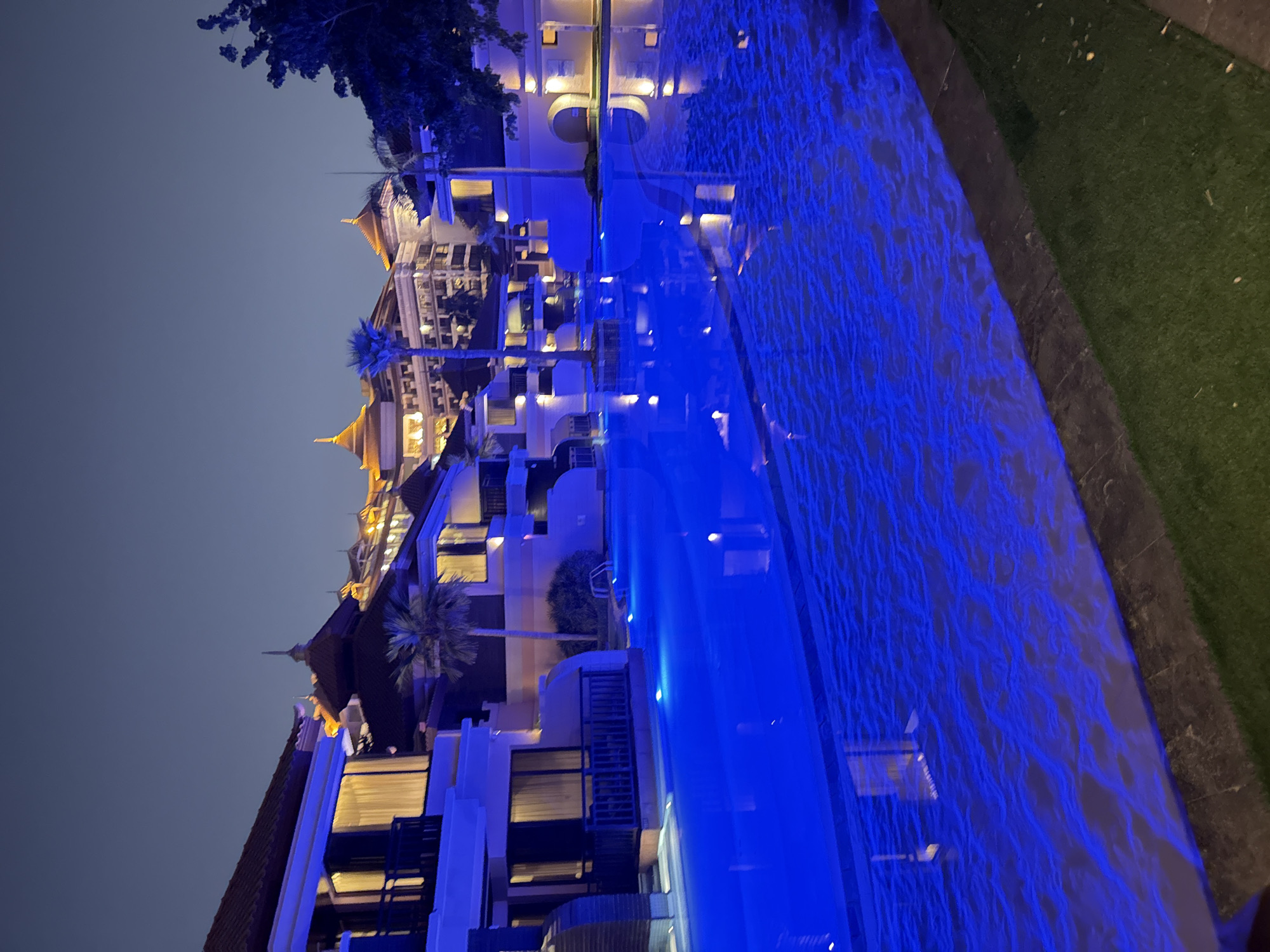 The image depicts a luxurious hotel complex at night, likely located in Dubai, United Arab Emirates, given the architectural style featuring distinctive domed roofs and intricate building designs. A large, illuminated swimming pool with vibrant blue underwater lighting dominates the foreground. The water's surface reflects the lights and the architecture, creating a mesmerizing effect. On the left and center of the image, modern hotel rooms or villas are visible, featuring balconies with dark railings and illuminated windows suggesting occupancy. Tropical palm trees are interspersed between the buildings, adding to the resort ambiance. In the background, a grand, multi-story hotel building with ornate golden-lit roofs and numerous windows stands prominently against the twilight sky. The overall scene conveys a sense of opulence, relaxation, and nighttime splendor typical of a high-end resort in Dubai.