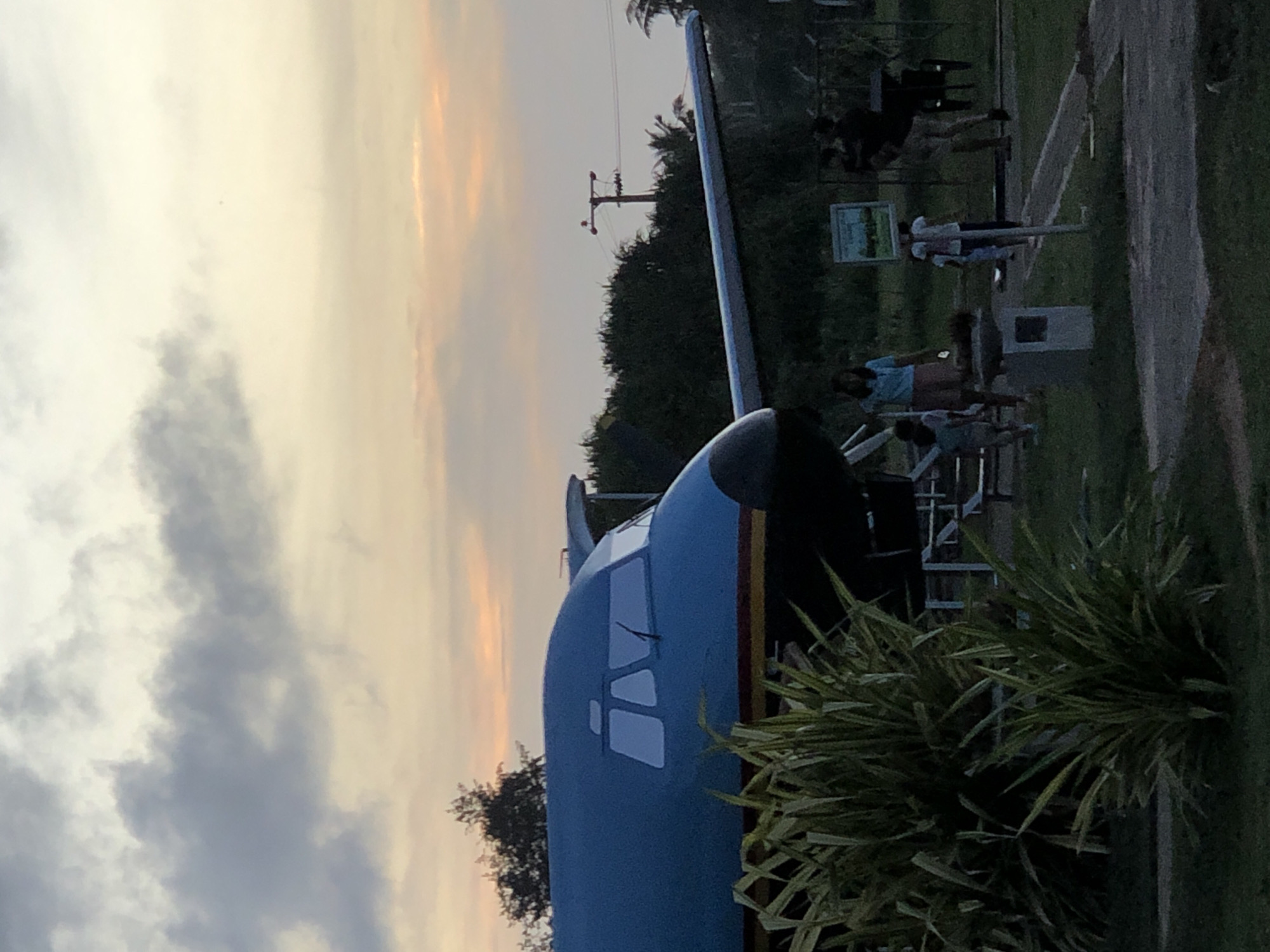The image depicts an outdoor scene at dusk, likely in Habaraduwa, Sri Lanka, featuring a grounded airplane with a blue fuselage and a red and yellow stripe. In the foreground, lush green foliage is prominent. Several individuals are present: a woman in a blue shirt and brown shorts, a child in a light blue shirt and shorts, another child in a white shirt and dark shorts, and two adults on the right side of the frame, one appearing to be a man with dark hair wearing a light-colored shirt and shorts. The background shows dense trees and a utility pole with power lines. The sky is characterized by dramatic clouds in shades of gray, yellow, and orange, indicative of sunset. The overall setting appears to be a park or a similar public space where the aircraft is on display.