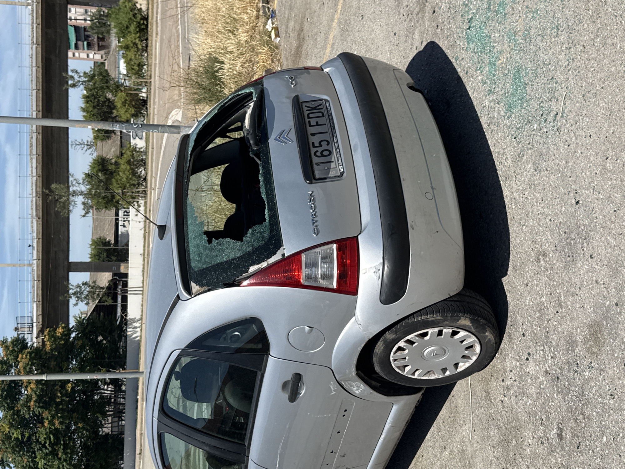 A silver Citroen C3 hatchback, license plate 1651FDK, is parked on an asphalt surface. The rear windshield is shattered, with glass fragments scattered on the ground. The car also shows signs of damage to the rear bumper and the body panel above the rear wheel, including what appear to be bullet holes or impacts on the driver's side rear door. Debris, including what looks like shattered glass and other litter, is present on the ground around the vehicle. In the background, a concrete overpass with a railing is visible, along with trees and urban buildings. The location is identified as L'Hospitalet de Llobregat, Spain. The scene suggests the car has been vandalized or involved in an incident causing significant damage.