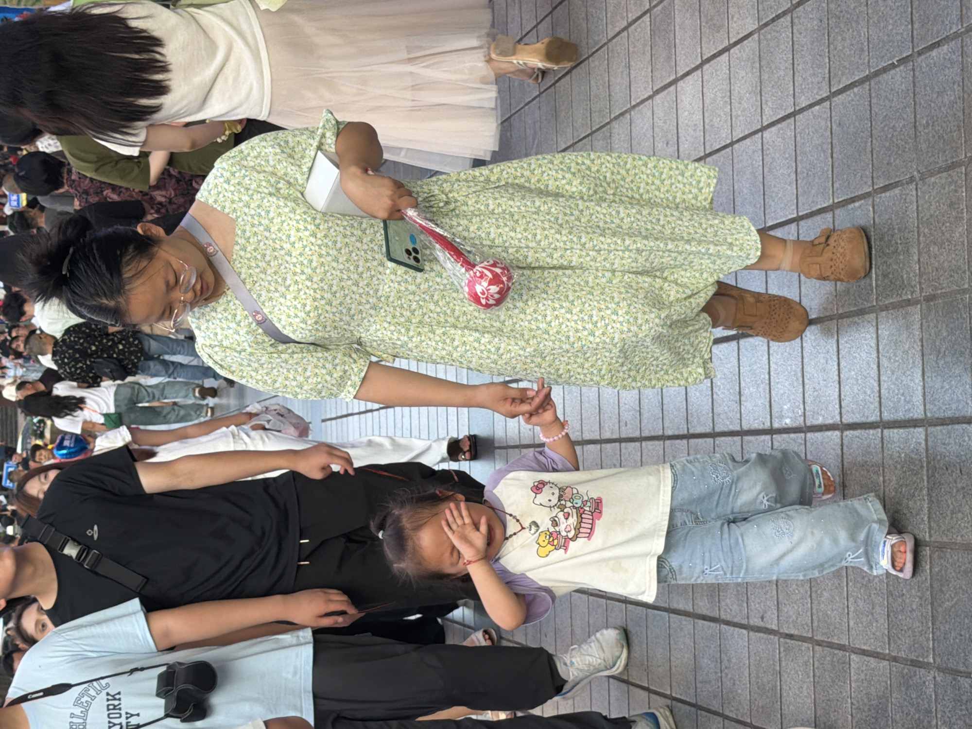 In Yuzhong District, China, a woman in a light green floral dress and tan loafers walks hand-in-hand with a young girl. The child, wearing a light purple and white t-shirt and light blue jeans, covers her eyes with her hand and appears to be crying. The woman holds a white box in her left arm and a red and white novelty item in a plastic bag in her right hand. They are walking on a tiled pedestrian walkway surrounded by other people. To their left, a person in a light blue t-shirt with a camera strap and a person in a black t-shirt with a crossbody bag are also visible. The environment is urban with a focus on the pedestrian area.