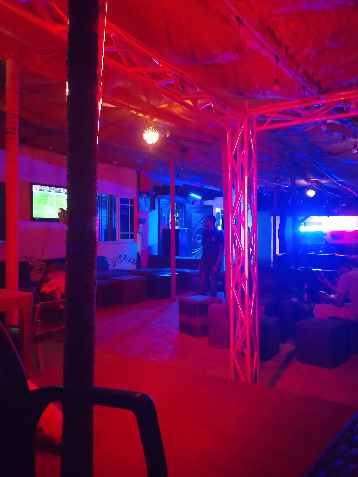 The image shows the interior of a bar or lounge in Yelwa, Nigeria. The lighting is primarily red and blue, creating a dim atmosphere. The walls are a light color, possibly white or beige. There are multiple low seating areas with cube-shaped stools and sofas, and a thatched roof overhead. In the background, a TV is showing a football match. There is a man standing near a sound system or DJ booth and another person seated. There is a window with musical notes and other posters and paintings on the walls. The floor appears to be made of bricks or tiles. The scene appears to be set up for entertainment and socializing.