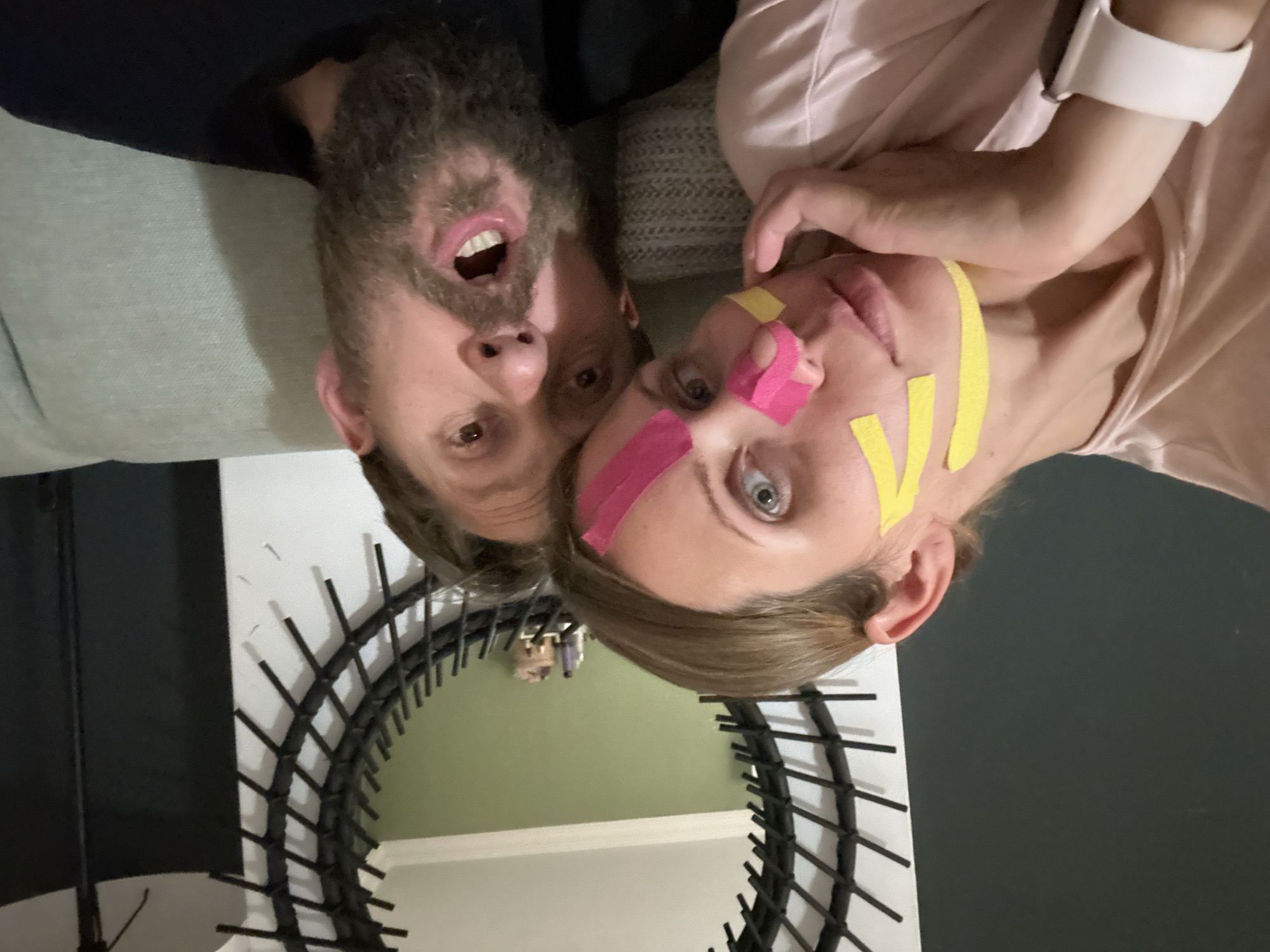 An indoor photograph, rotated 180 degrees, captures a man and a woman in an unconventional, playful pose. The man, positioned on the left, has a full beard and is wearing a dark top. His mouth is wide open, revealing his teeth, and his eyes are looking towards the viewer. The woman, on the right, wears a light-colored top and has light brown hair. Her face is adorned with pink and yellow kinesiology tape applied in various strips, and her eyes are wide open, looking directly forward. She has a white smartwatch on her left wrist.
Both individuals appear to be lying on a light grey sofa or couch, which forms part of the background behind them. Below their heads, a large, dark-framed decorative mirror with a spiky, radial design is prominent. The mirror reflects parts of the room, including an olive-green wall and a white wall, suggesting a decorated interior. A black, upright object, possibly a stand or an umbrella, is partially visible on the far left edge of the frame, near the mirror. The overall mood of the image is quirky and lighthearted, perhaps a moment of humor or an artistic selfie. There is no visible text in the image. The location context provided is Fethiye, Turkey.