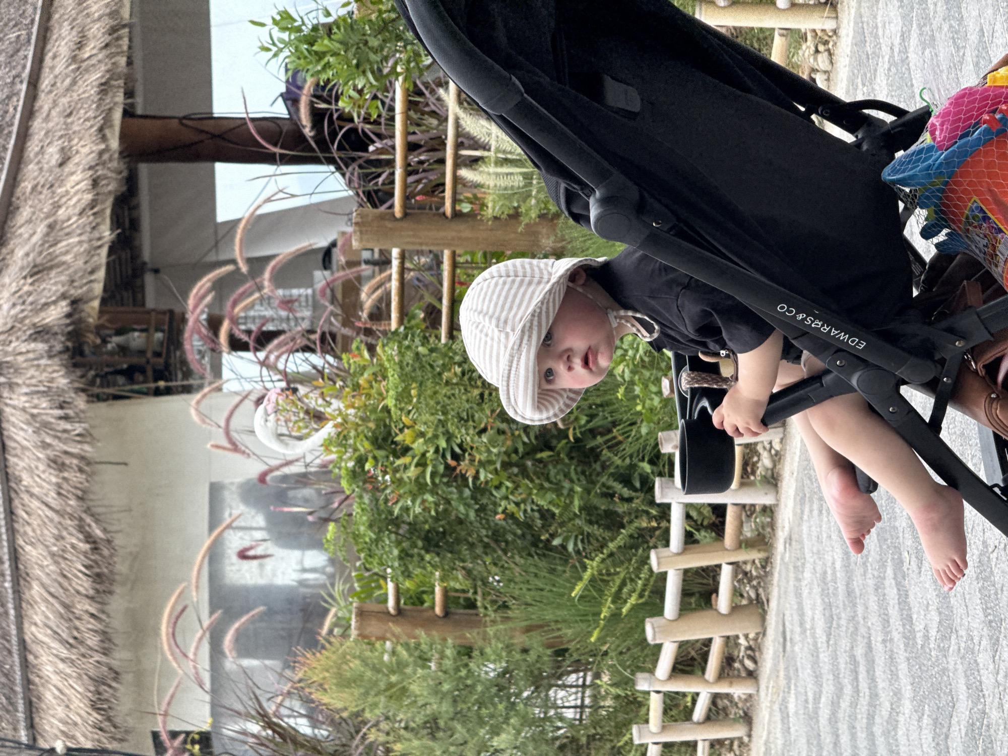 A young child, wearing a striped sun hat and a black shirt, is sitting in a black stroller. The child has fair skin and light-colored eyes, looking forward with a neutral expression. The stroller is positioned on a paved walkway, with a wooden fence and lush greenery in the background. Behind the fence, a building with a thatched roof is visible. The image appears to be taken outdoors, possibly in a park or garden area, with soft lighting. The child's bare feet are visible, resting on the stroller's footrest. A cup holder is attached to the stroller, and a net bag filled with colorful toys is hanging from the bottom. The scene suggests a leisurely outing in a tropical environment, potentially in Ko Samui District, Thailand, given the architectural style and vegetation.