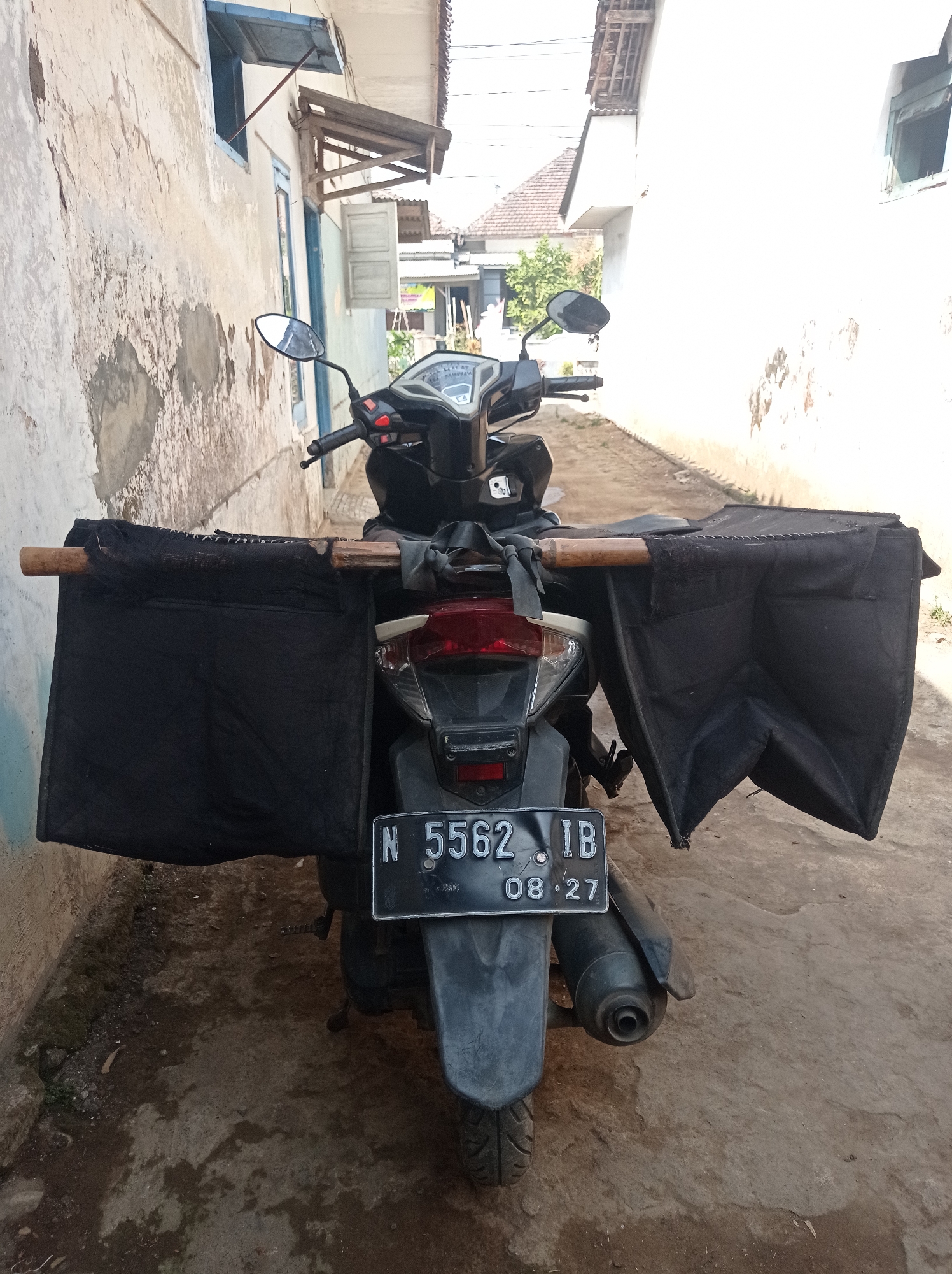 An outdoor, eye-level, medium shot captures the rear of a black motorcycle parked in what appears to be a narrow alleyway or street in Purwosekar, Indonesia. The motorcycle is equipped with two large, black fabric bags attached to a wooden pole extending from the rear. The bags are draped over the sides and hang down, creating a wide, almost wing-like appearance. A license plate, with the characters "N 5562 IB" and the date "08.27", is clearly visible on the rear fender. The motorcycle's taillight and exhaust pipe are also prominent. The environment consists of textured ground, likely concrete or dirt, with patches of dampness. To the left, a weathered, cream-colored building with peeling paint and a blue-trimmed window stands alongside the alley. To the right, a white building with a damaged facade and a window opening is visible. In the background, a glimpse of rooftops and greenery suggests a residential or rural setting. The scene is illuminated by natural daylight, casting shadows. There are no people present in the image.