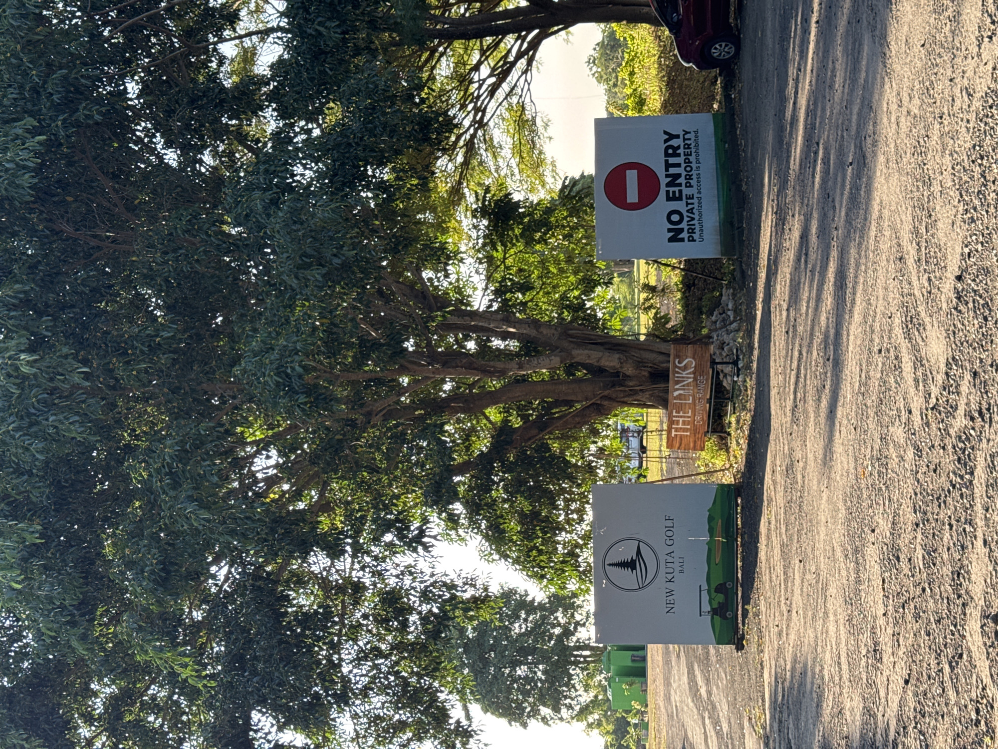 The image shows an entrance to New Kuta Golf in Bali, Indonesia. The scene is a paved road flanked by signs and trees. A sign for "New Kuta Golf Bali" is visible on the left side of the road. Above it is a wooden sign for "The Links Driving Range". On the right side of the road is a "NO ENTRY PRIVATE PROPERTY" sign. Lush trees surround the signs, with their branches and foliage filling much of the frame. A glimpse of a car is seen at the top of the image on the right, near the "NO ENTRY" sign. The road appears to lead towards the golf course or driving range in Kuta Selatan, Indonesia.
