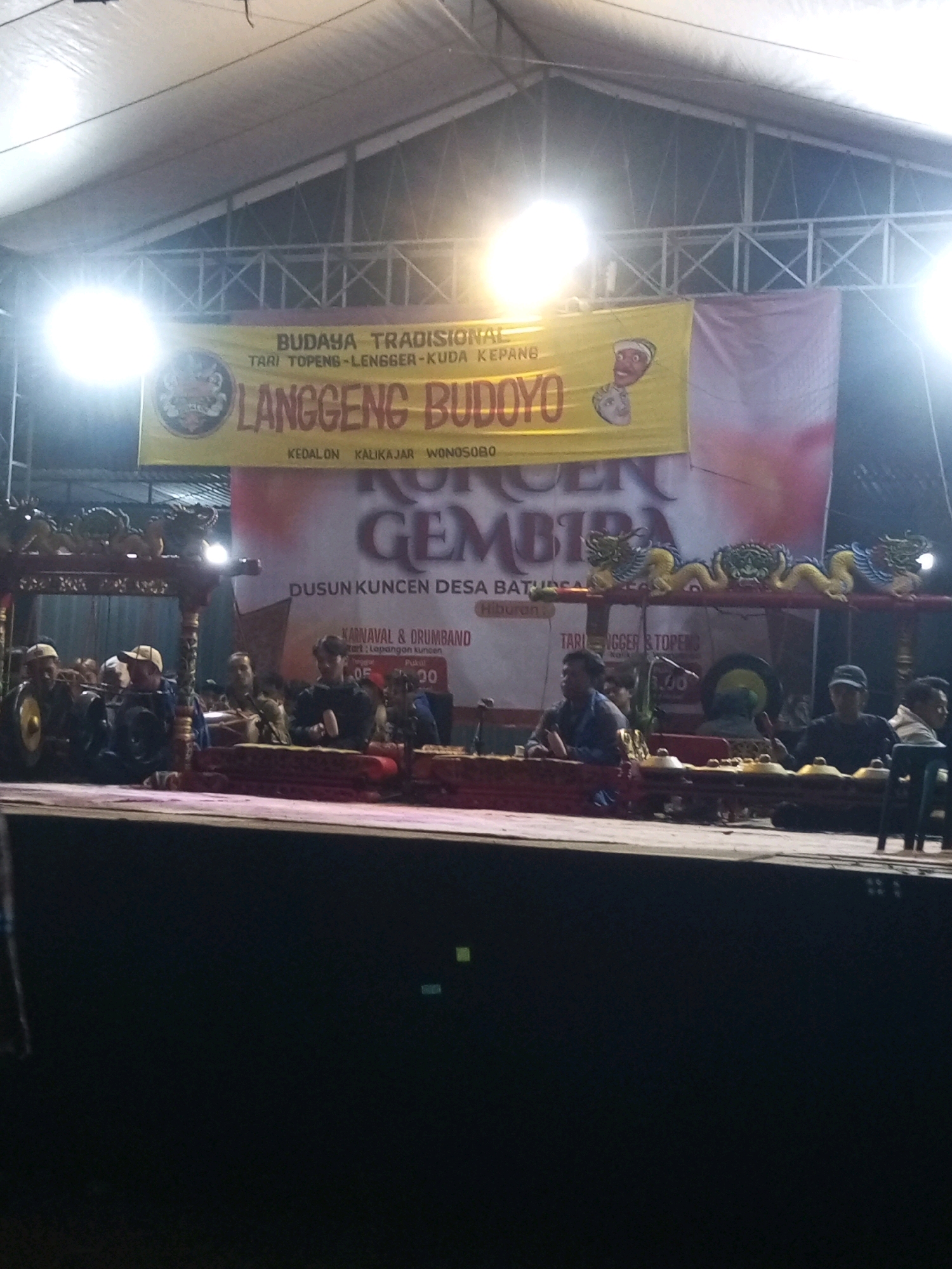 In the city of Ngadikerso, Indonesia, an outdoor stage is set up under a large white tent. The stage features traditional Indonesian musical instruments, including a gamelan. Several people are seated on the stage, some appearing to be musicians. A banner behind them reads "BUDAYA TRADISIONAL" (Traditional Culture) and displays the name "LANGGENG BUDOYO" and "KULUK GEMBIRA". Below the banner, smaller text indicates "DUSUN KUNCEN DESA BATUSONGO" (Kuncen Hamlet, Batusongo Village) and "KARNAVAL & DRUMBAND". The event appears to be a cultural performance or festival. The lighting suggests it is evening or nighttime.