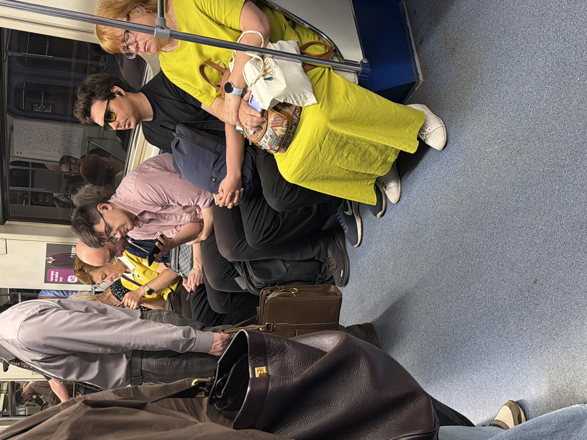 The image depicts the interior of a Moscow Metro train carriage. Several passengers are seated, facing the camera. In the foreground, a person's torso and arm are visible, holding a dark brown leather bag. To their right, a man in a pink shirt is looking at his phone, wearing earbuds. Next to him, a man in a black t-shirt and yellow sunglasses is also wearing earbuds. Further to the right, a woman in a bright yellow dress is seated, holding a small patterned bag and a white bag. She is wearing glasses and earbuds. Other passengers are visible in the background, some looking at their phones. The train carriage has blue seating and a light blue floor. Advertisements are visible on the wall behind the passengers, one featuring text in Russian. The overall scene is one of daily commute within the Moscow Metro.