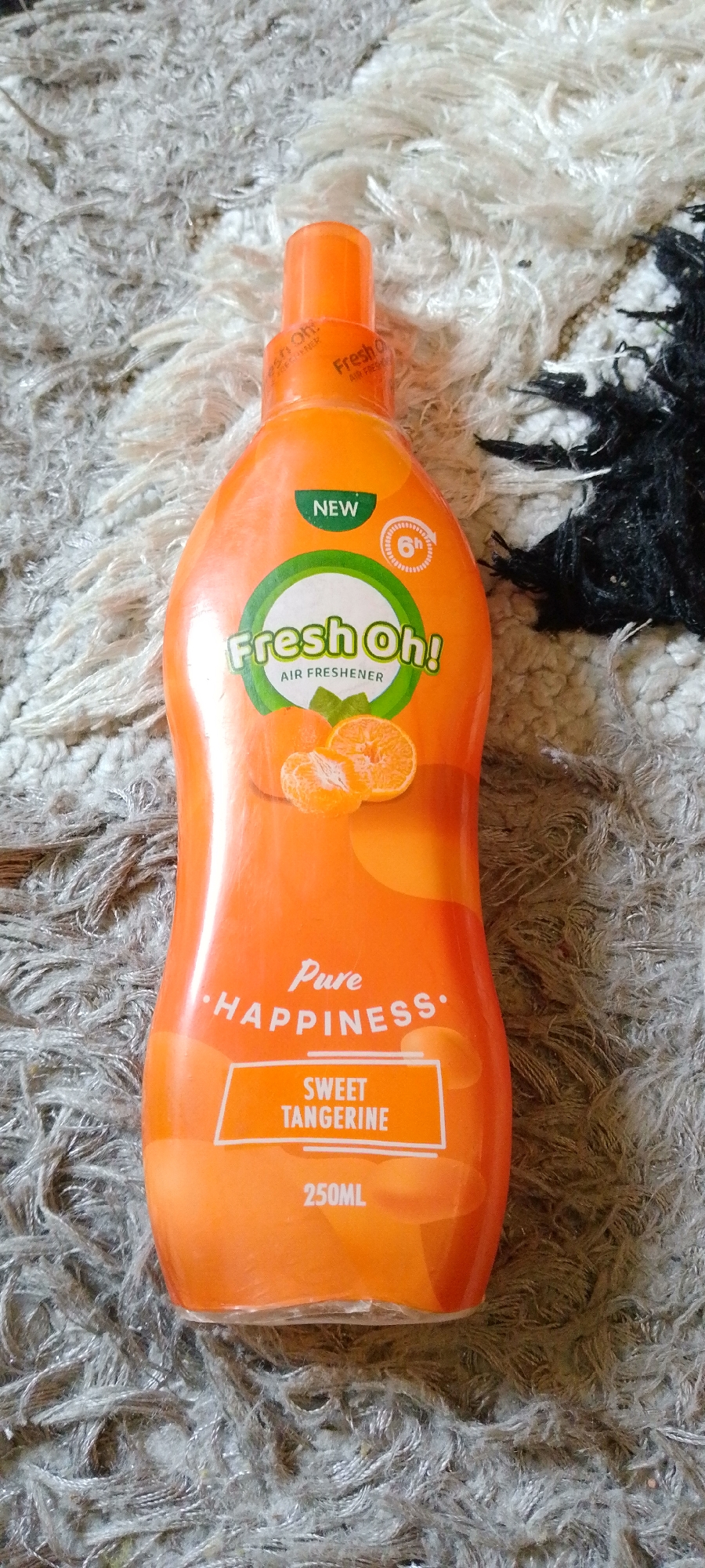 The image shows an orange plastic bottle of "Fresh Oh! Pure Happiness Sweet Tangerine Air Freshener." The bottle has a bright orange cap and the label is predominantly white and green with orange accents. The label features the brand name "Fresh Oh!" in large green lettering, with smaller text below indicating "AIR FRESHENER." There is also a "NEW" tag and a circular graphic with "6h" suggesting a 6-hour duration. Images of sliced tangerines are depicted on the label. Below this, "Pure HAPPINESS" is written in a script font, followed by "SWEET TANGERINE" in a white box. The bottle is labeled with a volume of "250ML". The object is placed on a textured surface that appears to be a rug with a mix of white and grey fibers, and some black fibers on the right side. The lighting suggests it is indoors, possibly daylight. No people are visible in the image. The location context provided is Jauro Yinu, Nigeria.