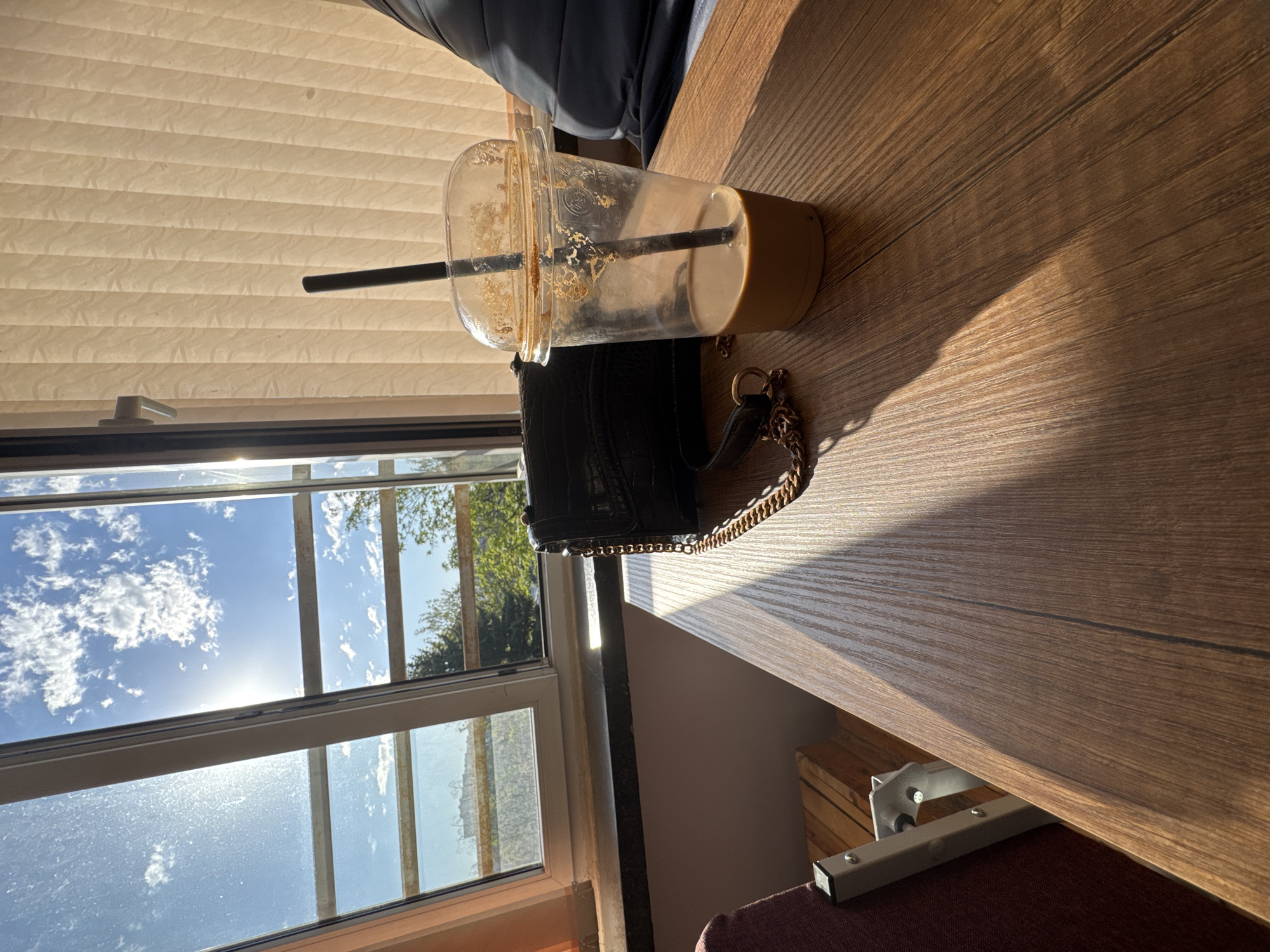 The image shows a scene inside a building, possibly a home or office, in what appears to be Sofia, Bulgaria, based on the architectural style visible through the window. The photo is taken at an angle, with a wooden desk surface dominating the foreground. On the desk, there is an empty plastic coffee cup with a black straw, suggesting it was recently used. Next to the cup is a small black purse with a gold chain strap. A window is visible to the left, revealing a bright blue sky with scattered white clouds. The sunlight streaming through the window casts a warm glow on the desk surface and the objects on it. A window blind is partially drawn in the upper left. A chair is partially visible behind the table.